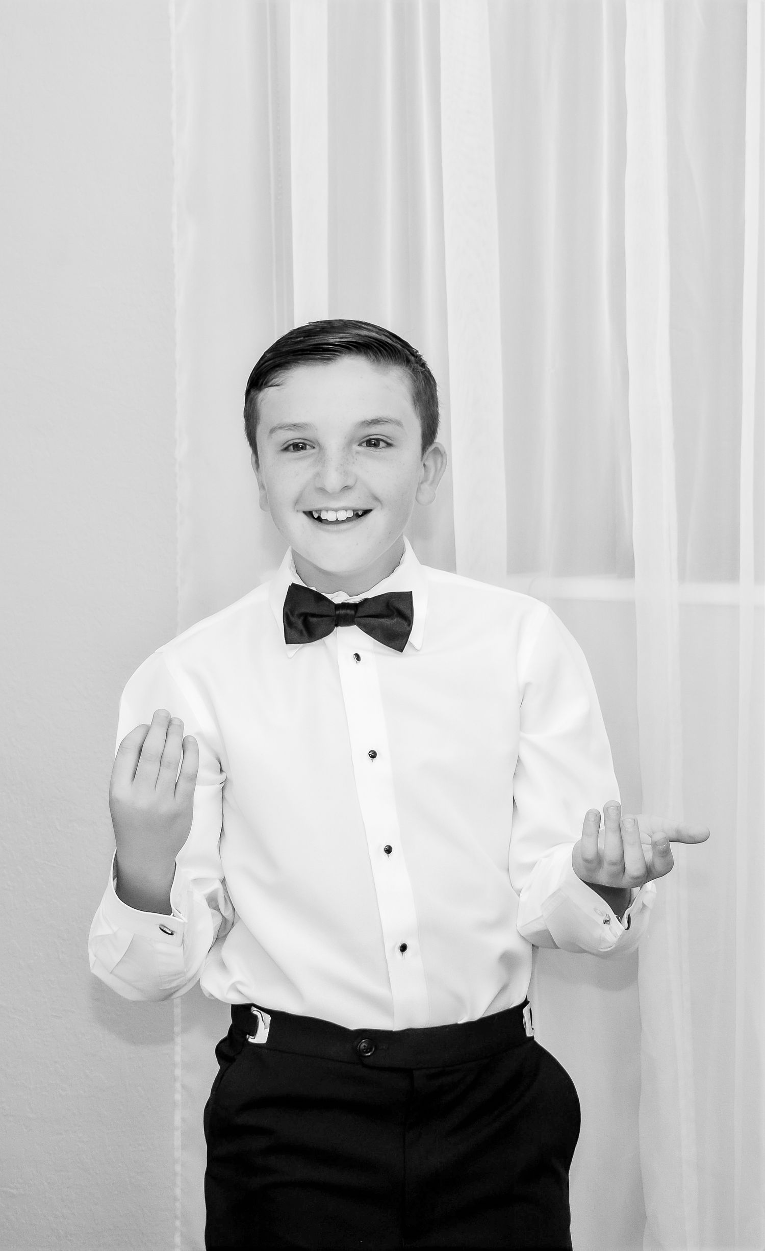 Young gentleman in formal attire with bow tie poses playfully against white backdrop.