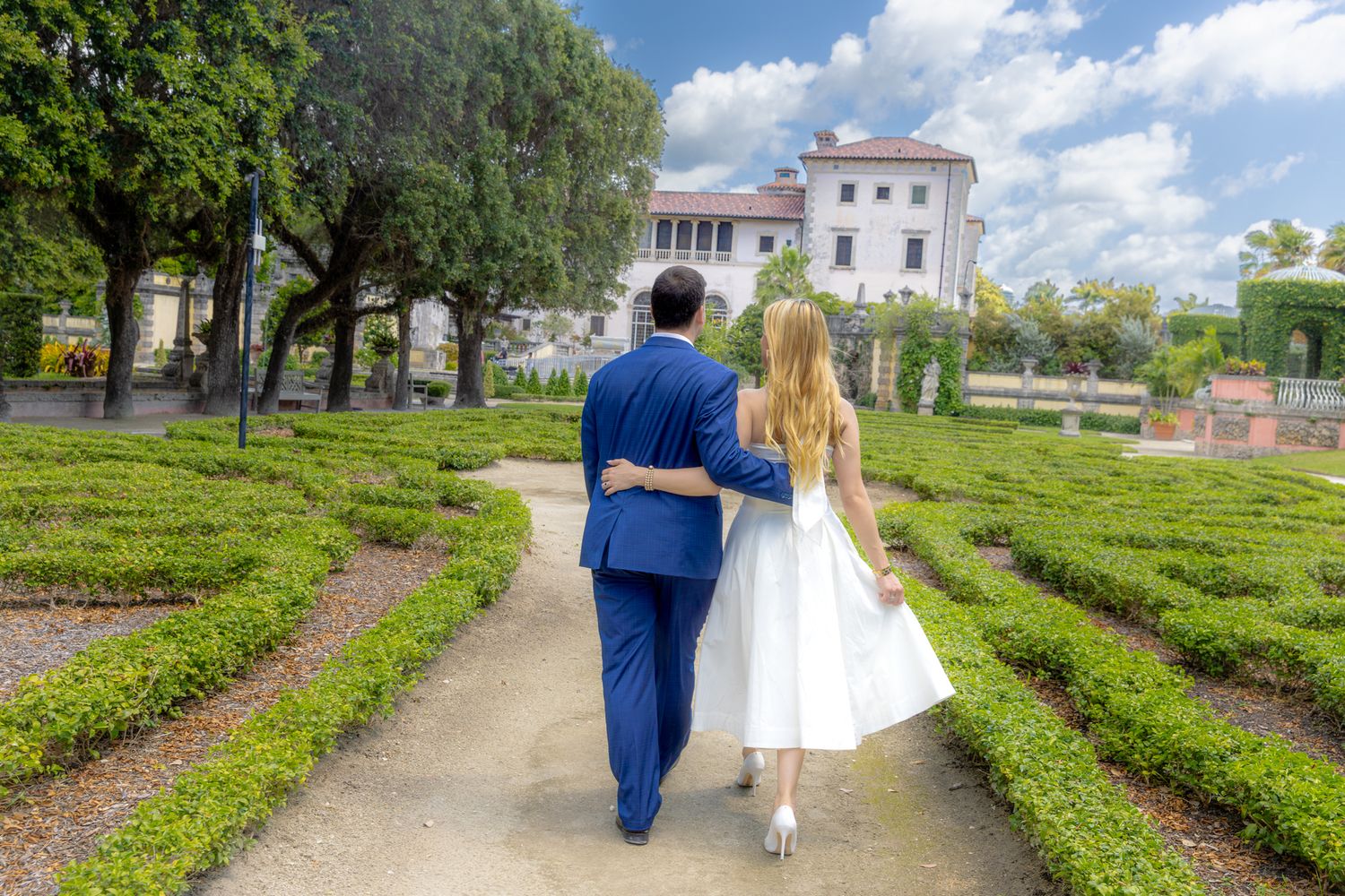 A couple in formal attire walks along a garden path towards a white mansion on a sunny day.