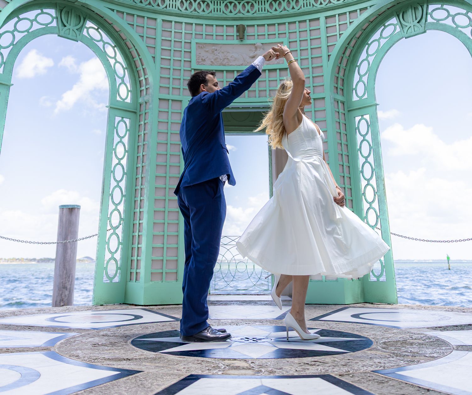 Couple dances together in a mint green gazebo overlooking the ocean on a bright sunny day.