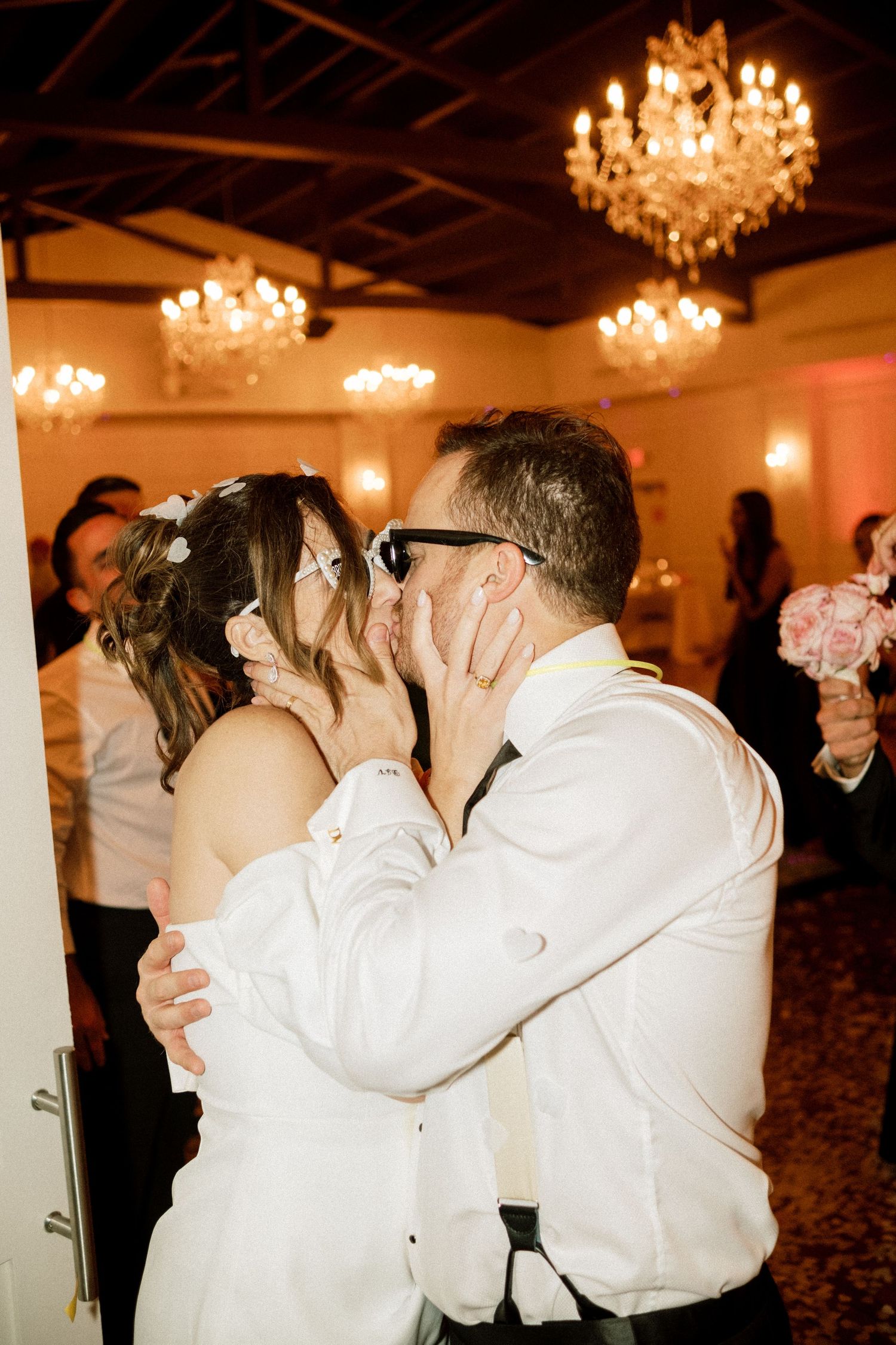 Wedding guests dance and embrace on an elegant ballroom dance floor under crystal chandeliers.