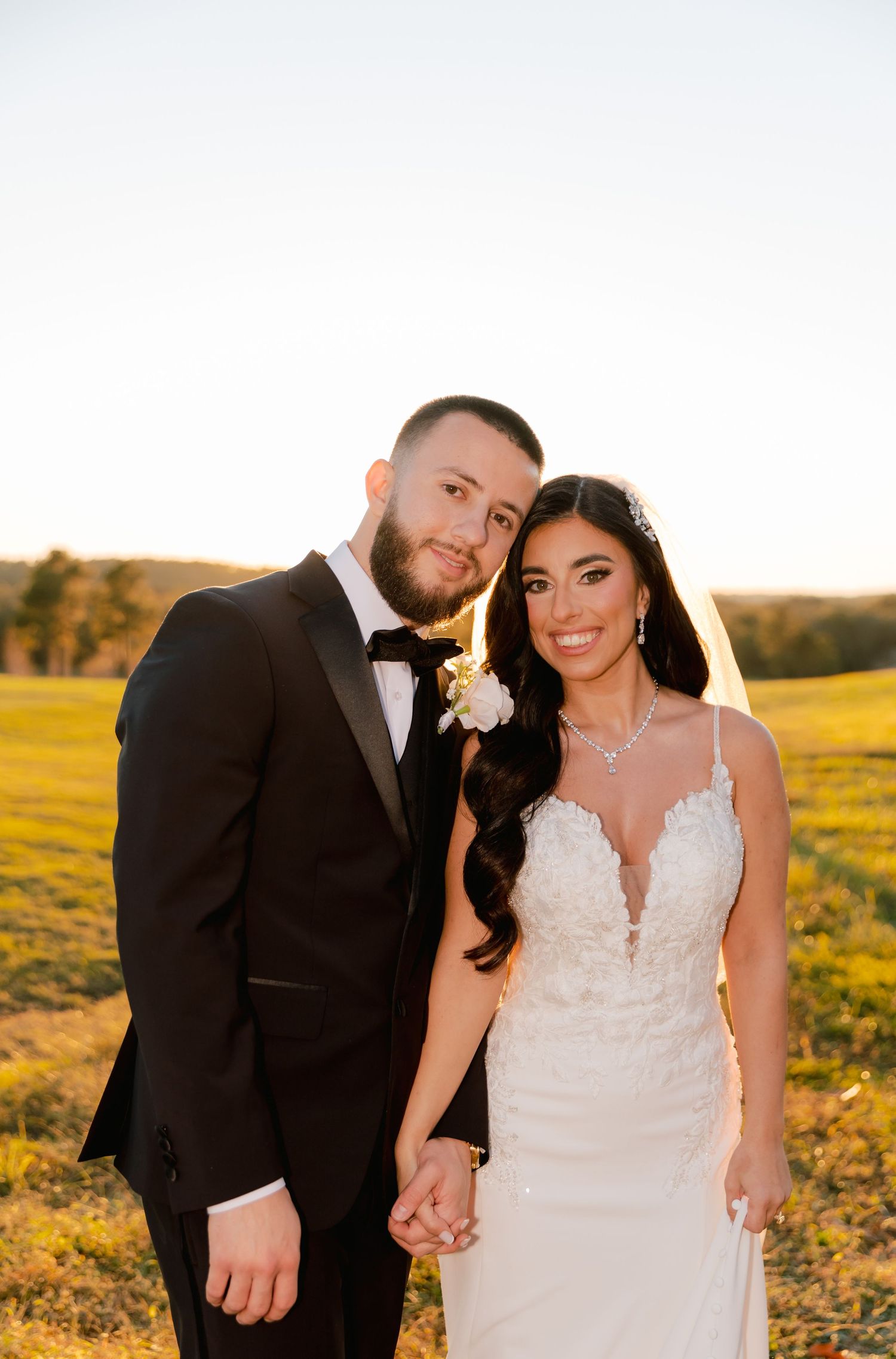 A couple poses together in formal wedding attire in a golden field during sunset.