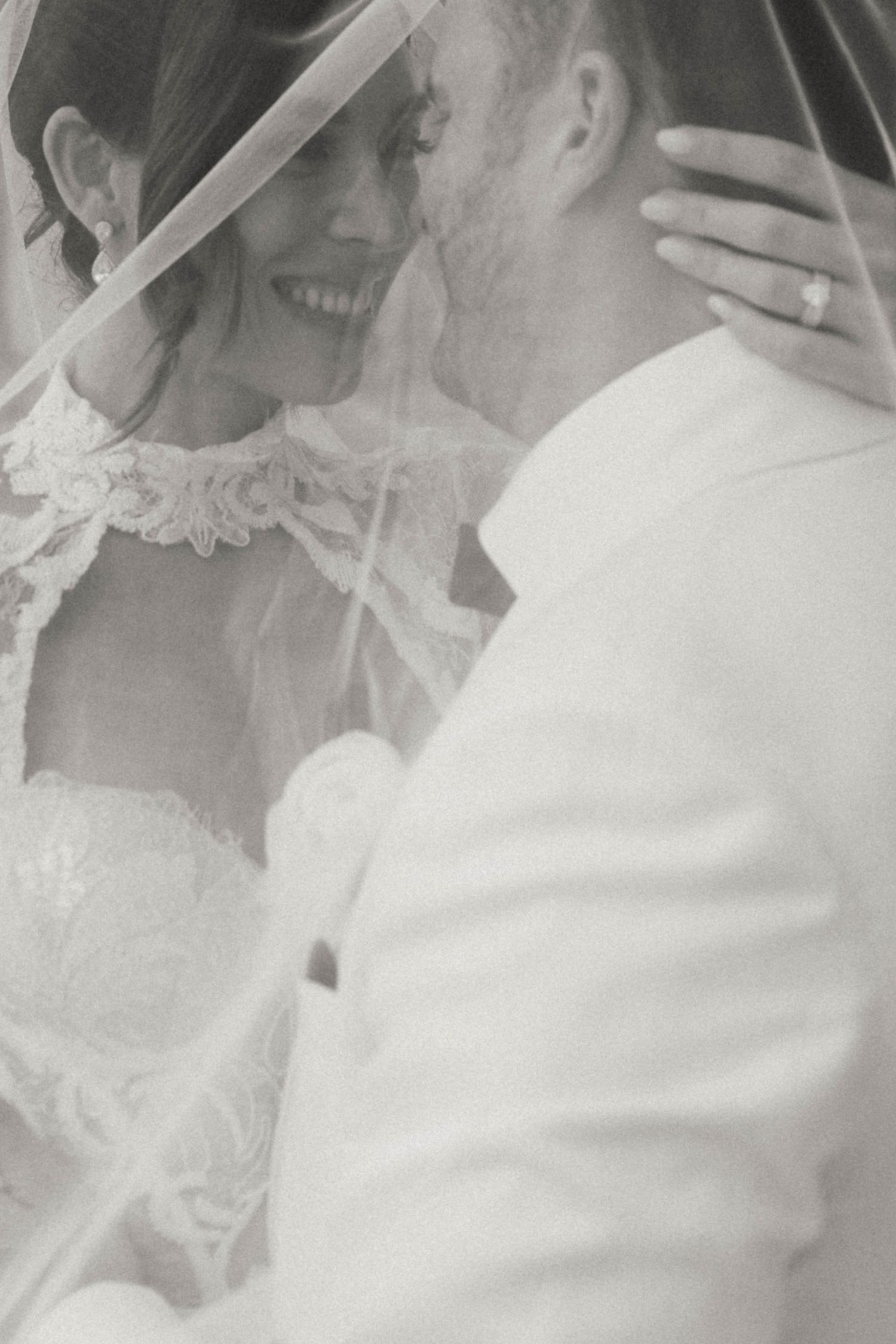 A tender black and white wedding photograph showing a couple embracing under a delicate lace veil.