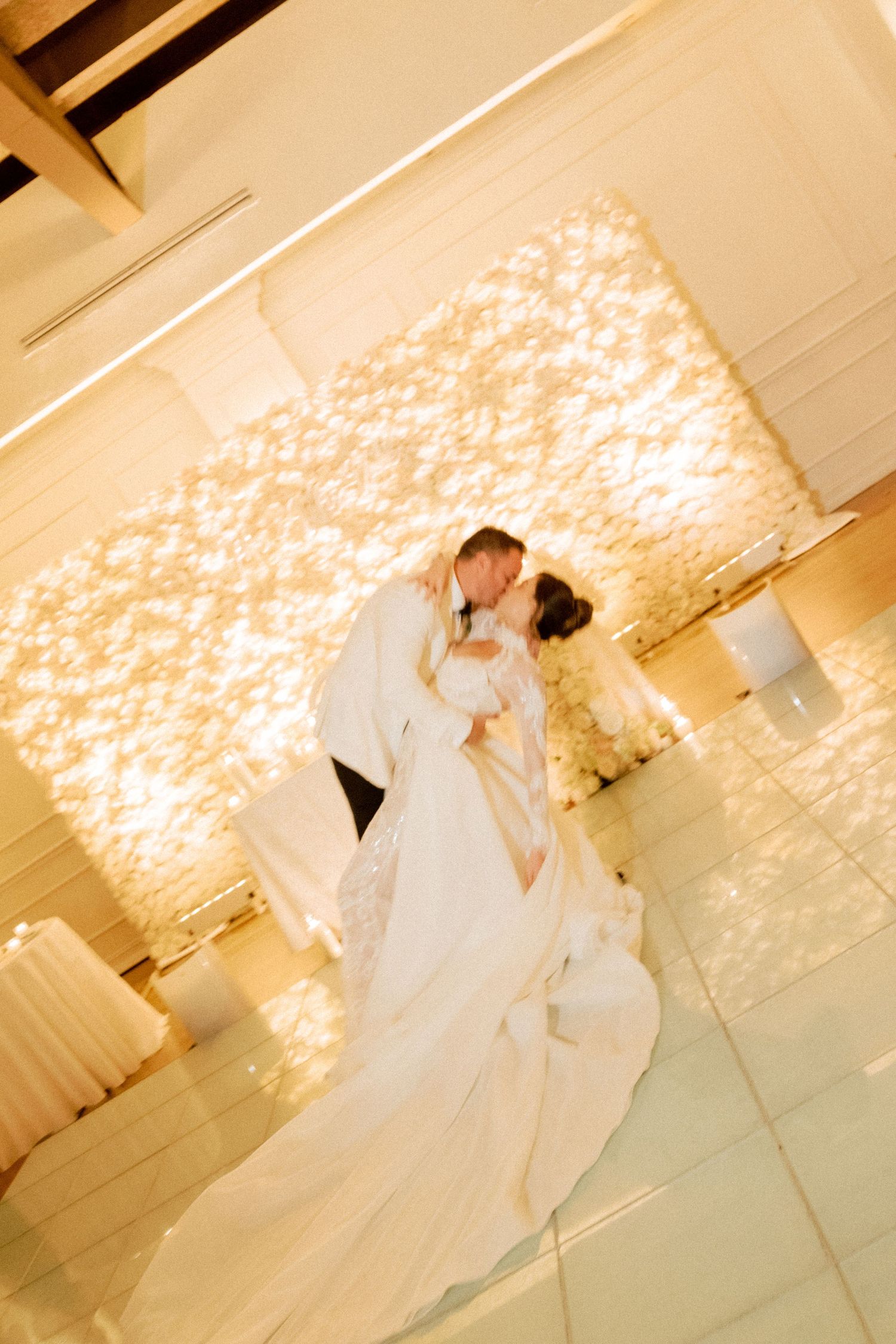 A couple in white attire shares a romantic dance moment on a white marble floor with illuminated wall patterns.