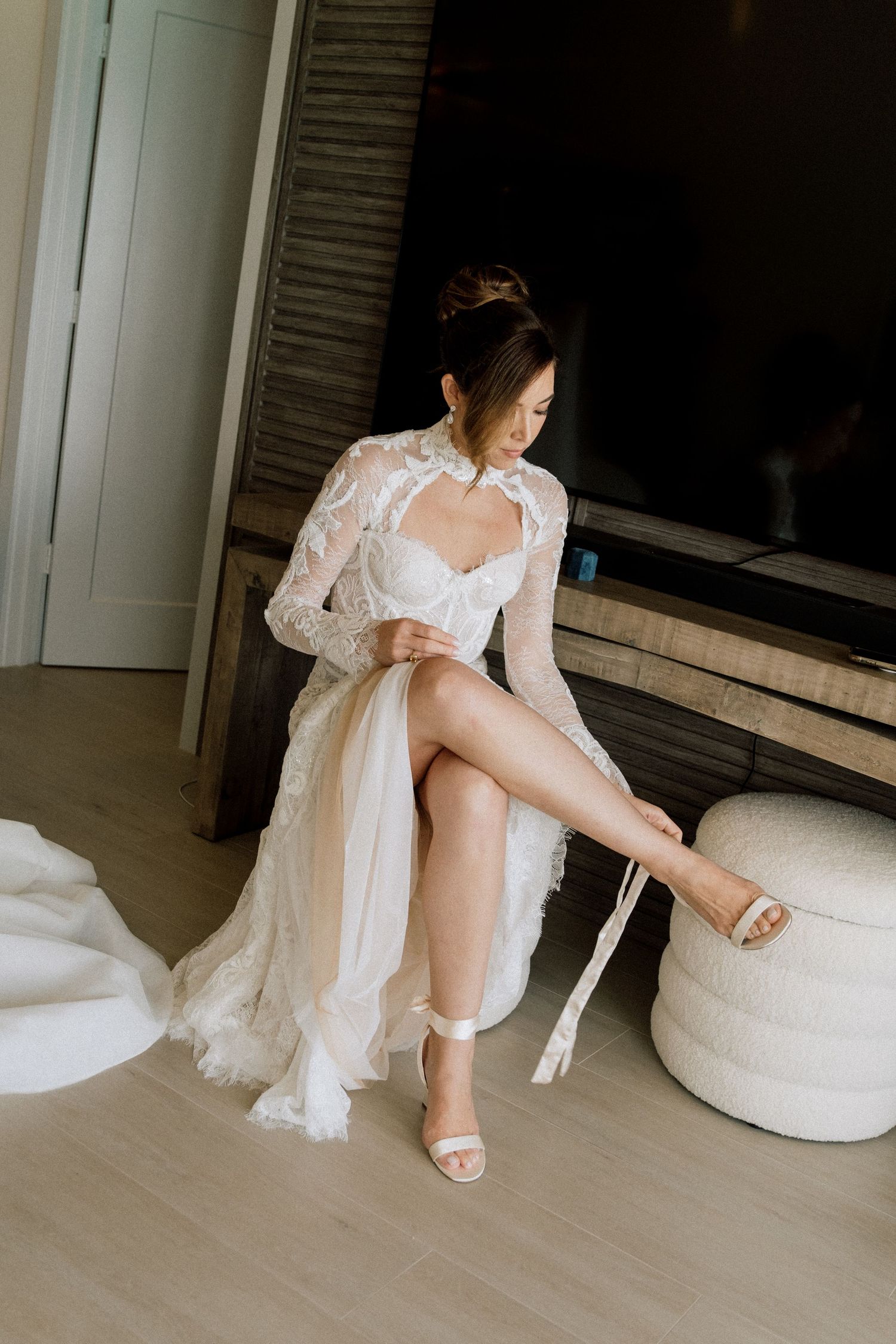 A bride in a white wedding dress sits on the floor while putting on elegant white heels.