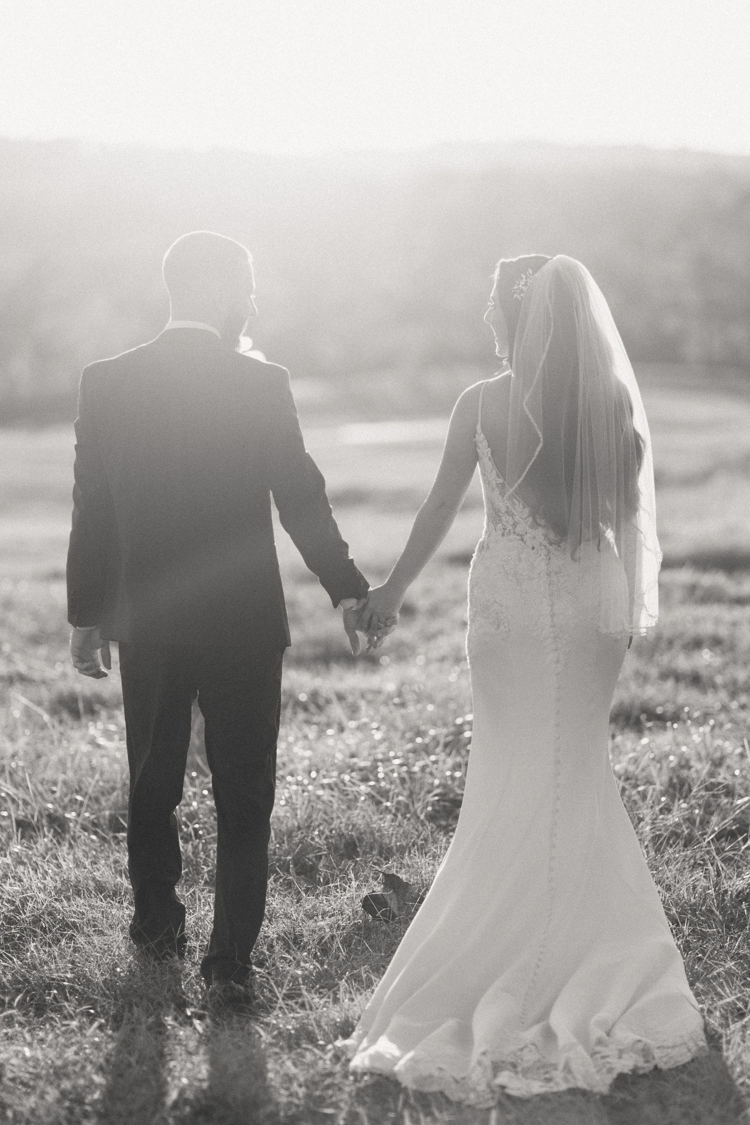 A bride and groom hold hands while walking together in a misty field at sunset, captured in black and white.