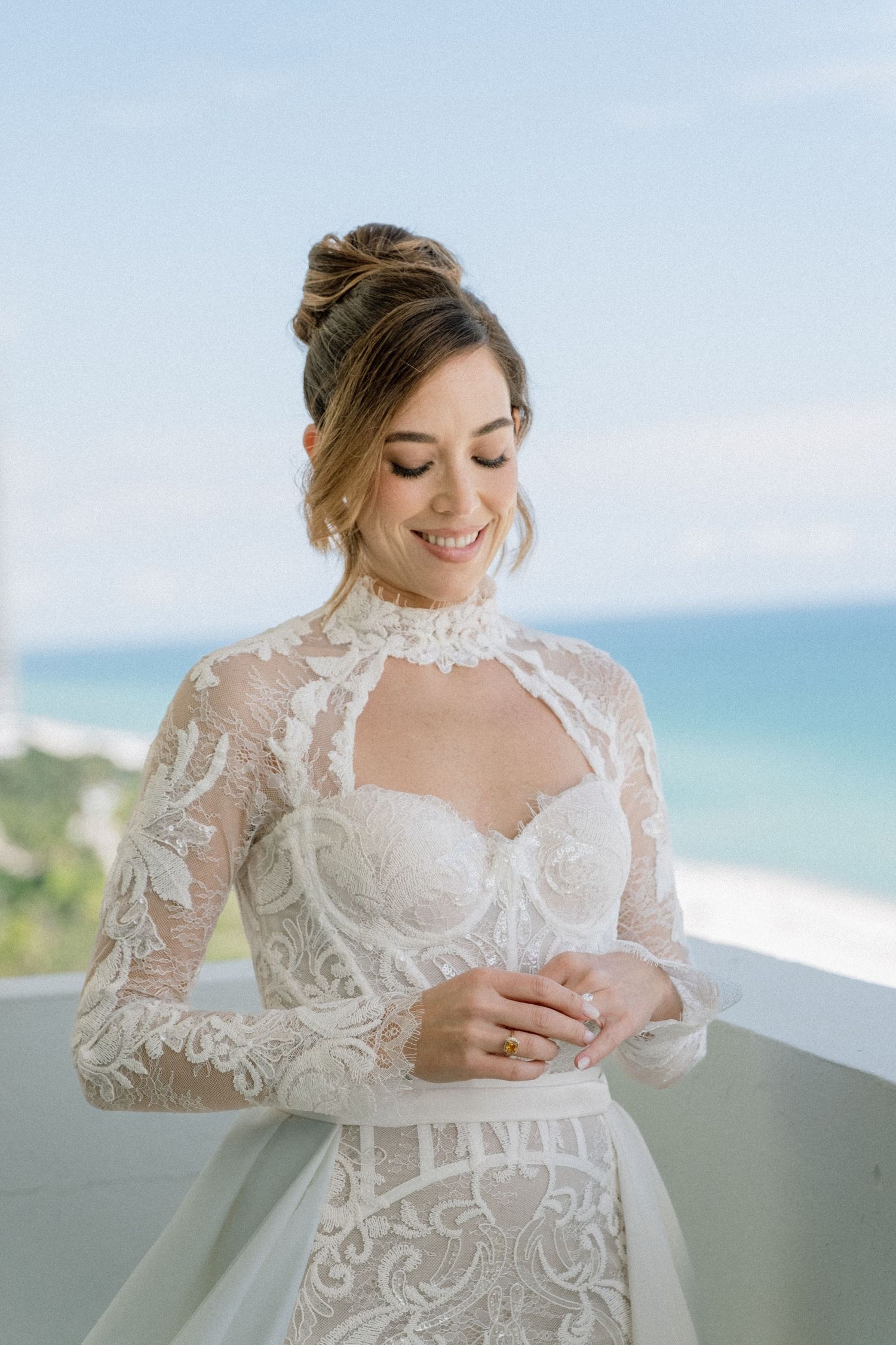 A bride in an elegant lace wedding dress with long sleeves reads a letter on a balcony overlooking the ocean.
