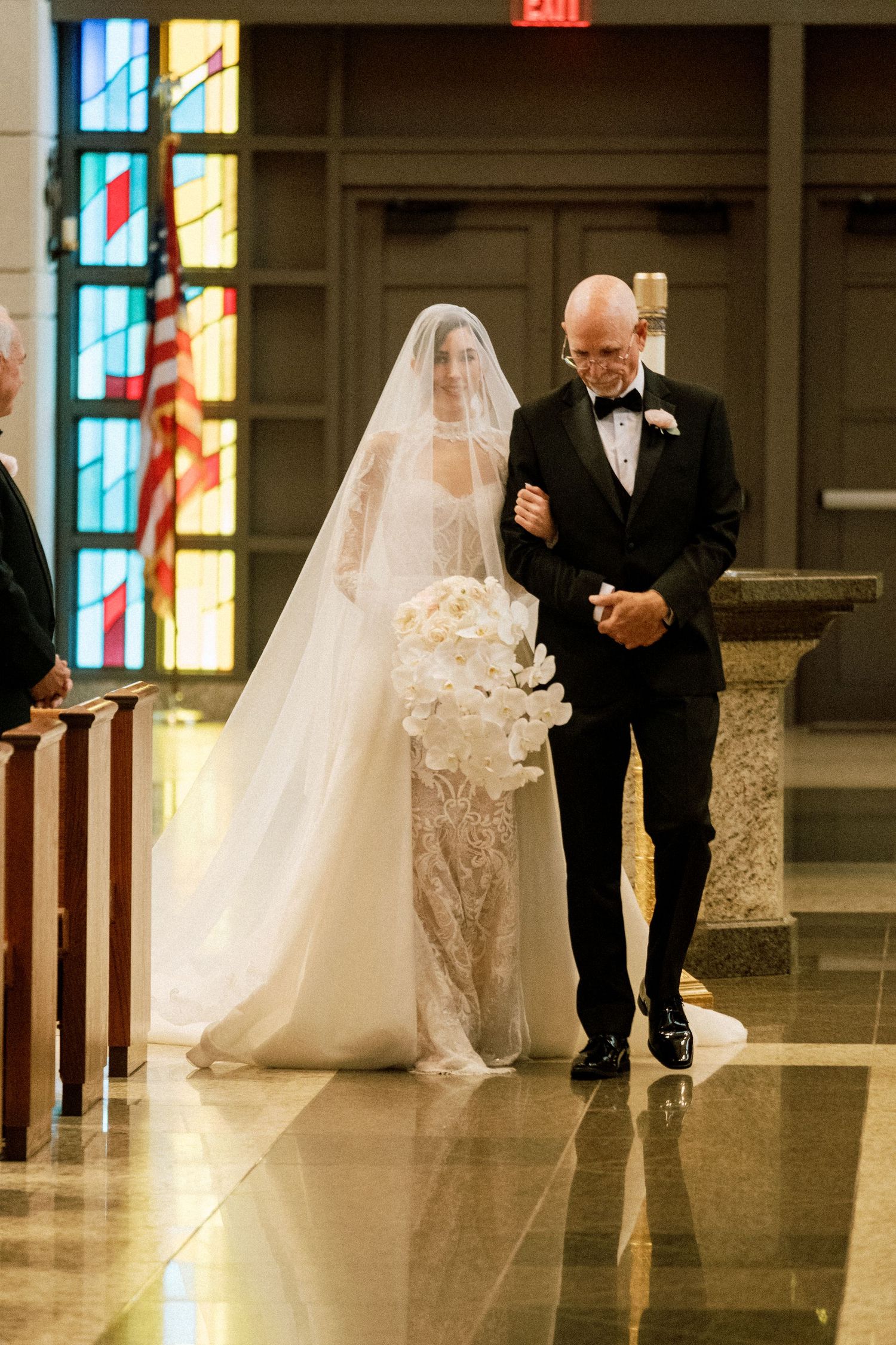A bride in a flowing white gown and veil walks down the aisle with an escort at a church wedding ceremony.