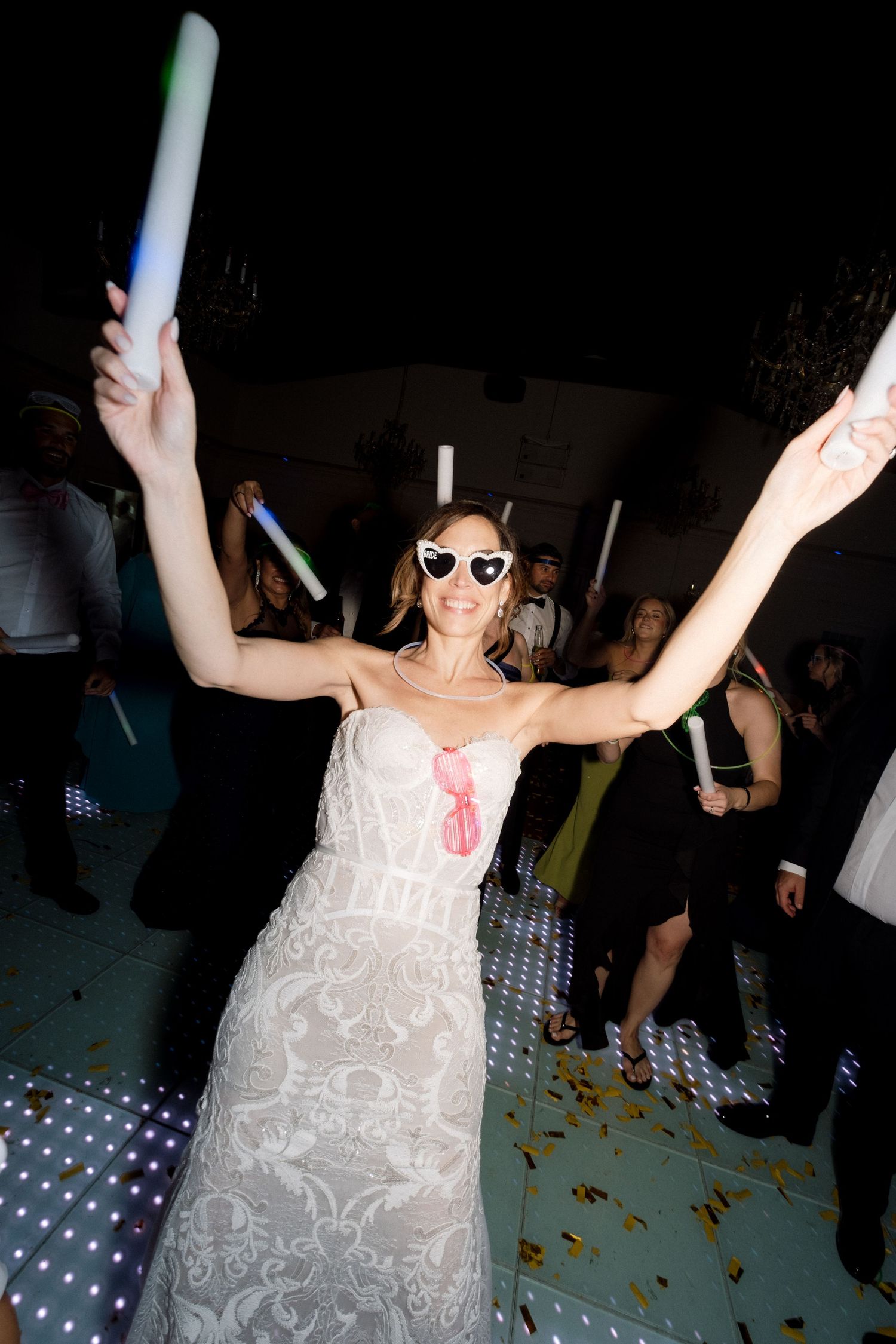 Party guest in white dress celebrating with glow sticks and sunglasses on the dance floor.