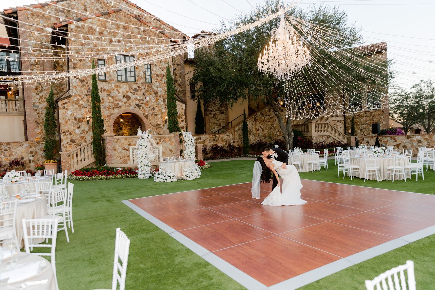A couple shares their first dance on a wooden dance floor at an outdoor wedding reception with stone walls and string lights.