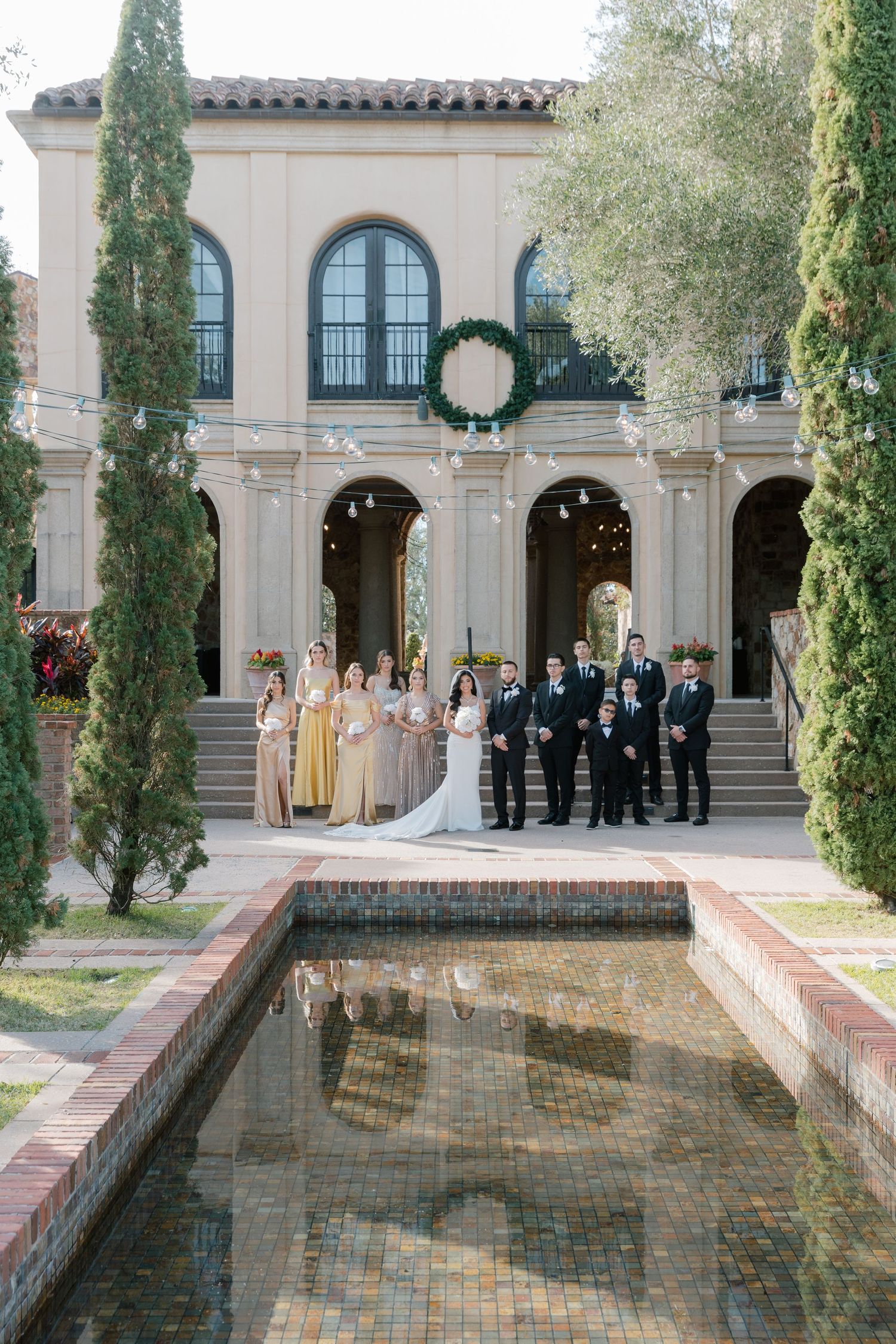 Wedding party stands elegantly arranged along a Mediterranean-style courtyard with reflecting pool and cypress trees.