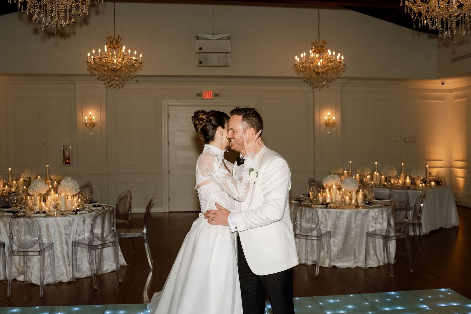 Elegant wedding couple dances in a grand ballroom with crystal chandeliers and white table settings.
