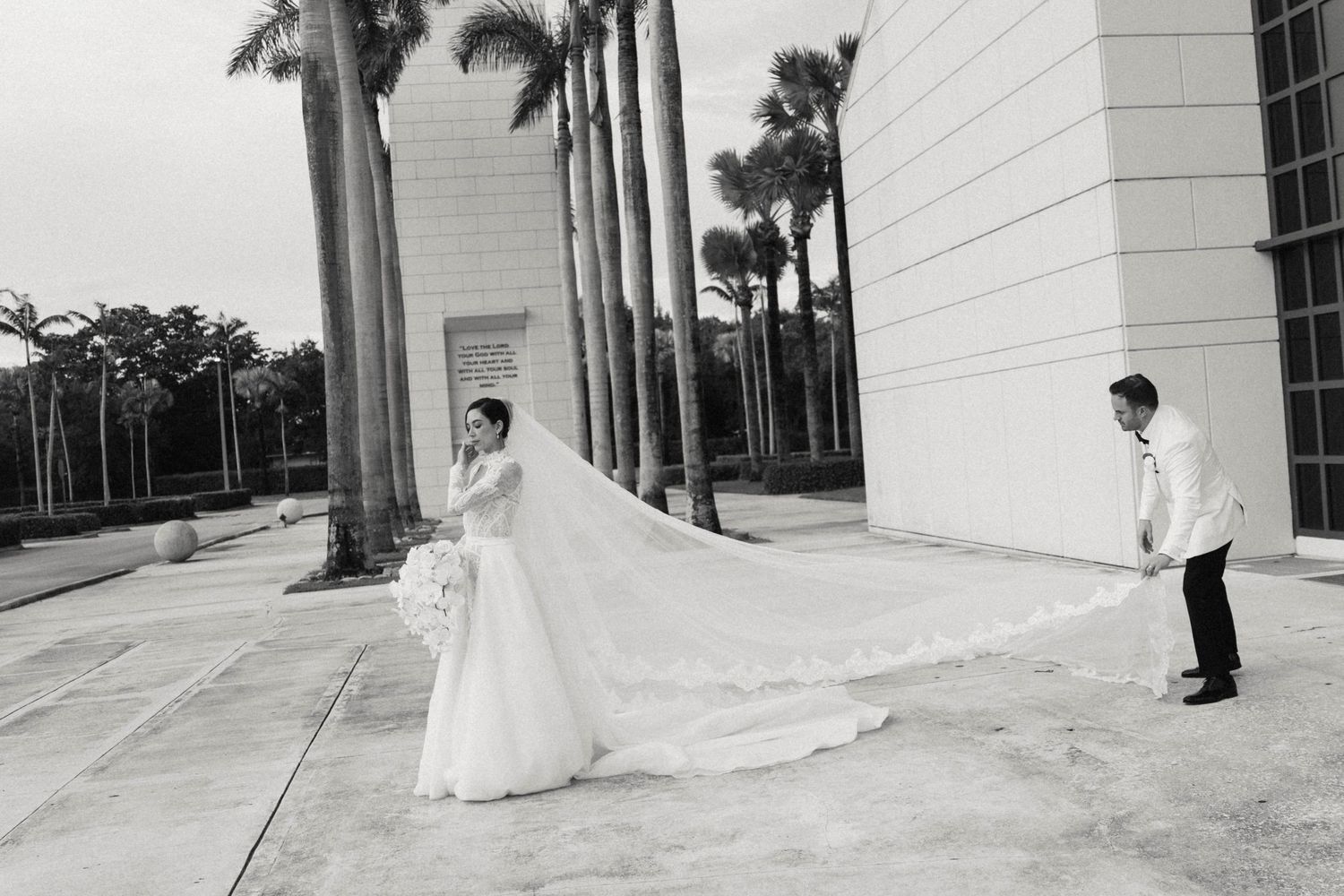 A dramatic black and white wedding photo with a flowing cathedral veil outside a modern building with palm trees.