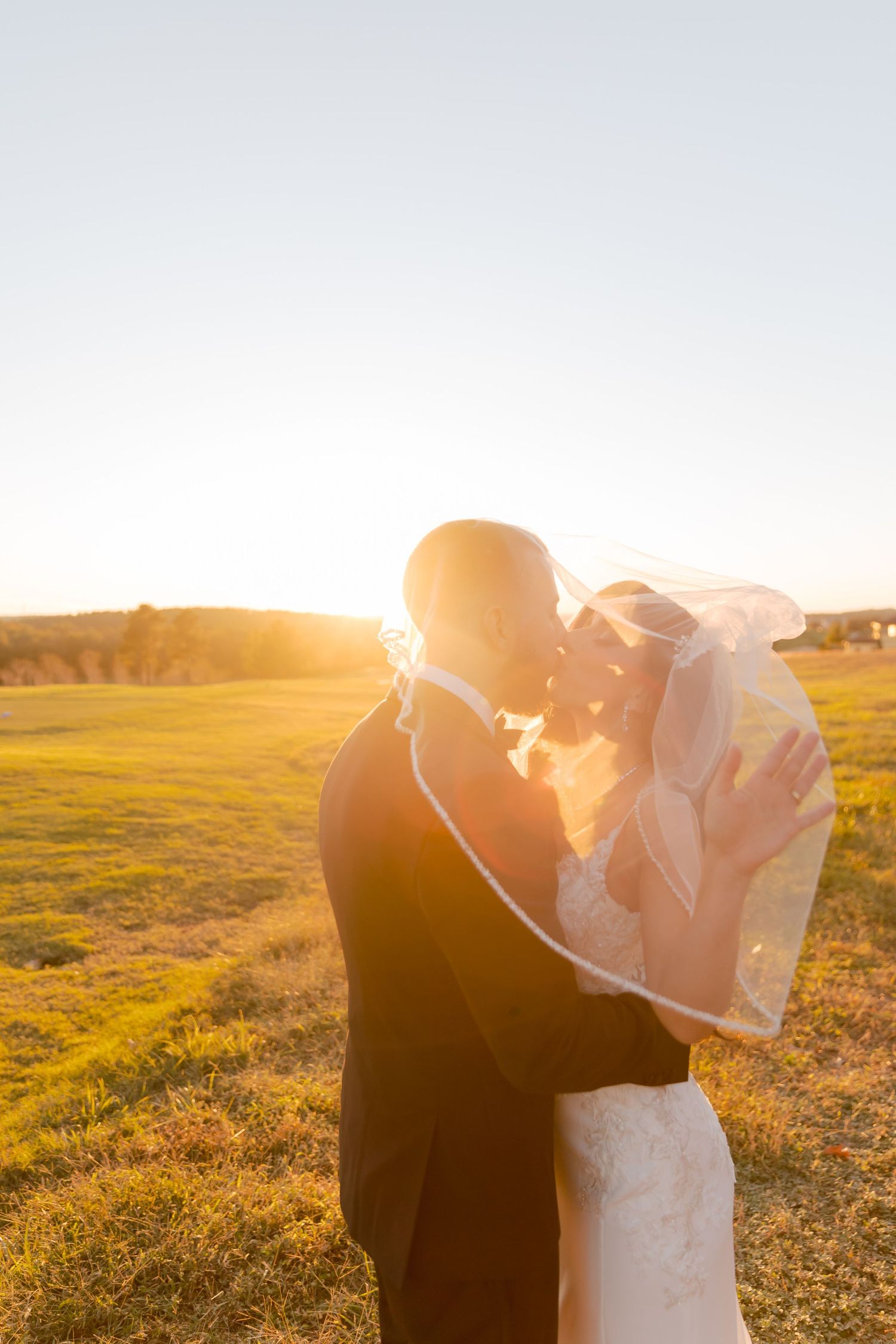 A bride and groom embrace during golden hour in a sunlit field at sunset while her veil flows in the breeze.
