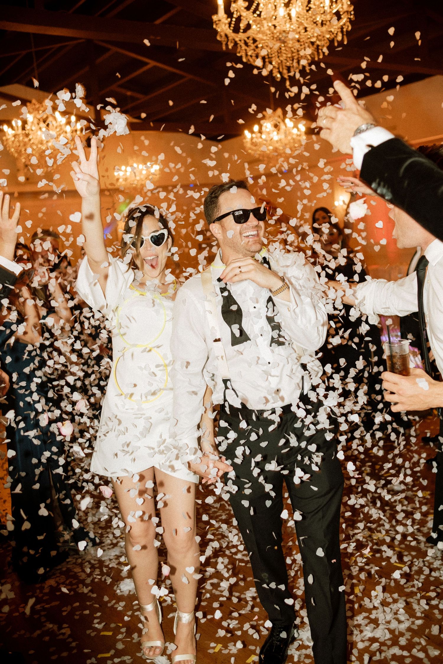 Wedding celebration with confetti falling over dancing guests under warm chandelier lighting.