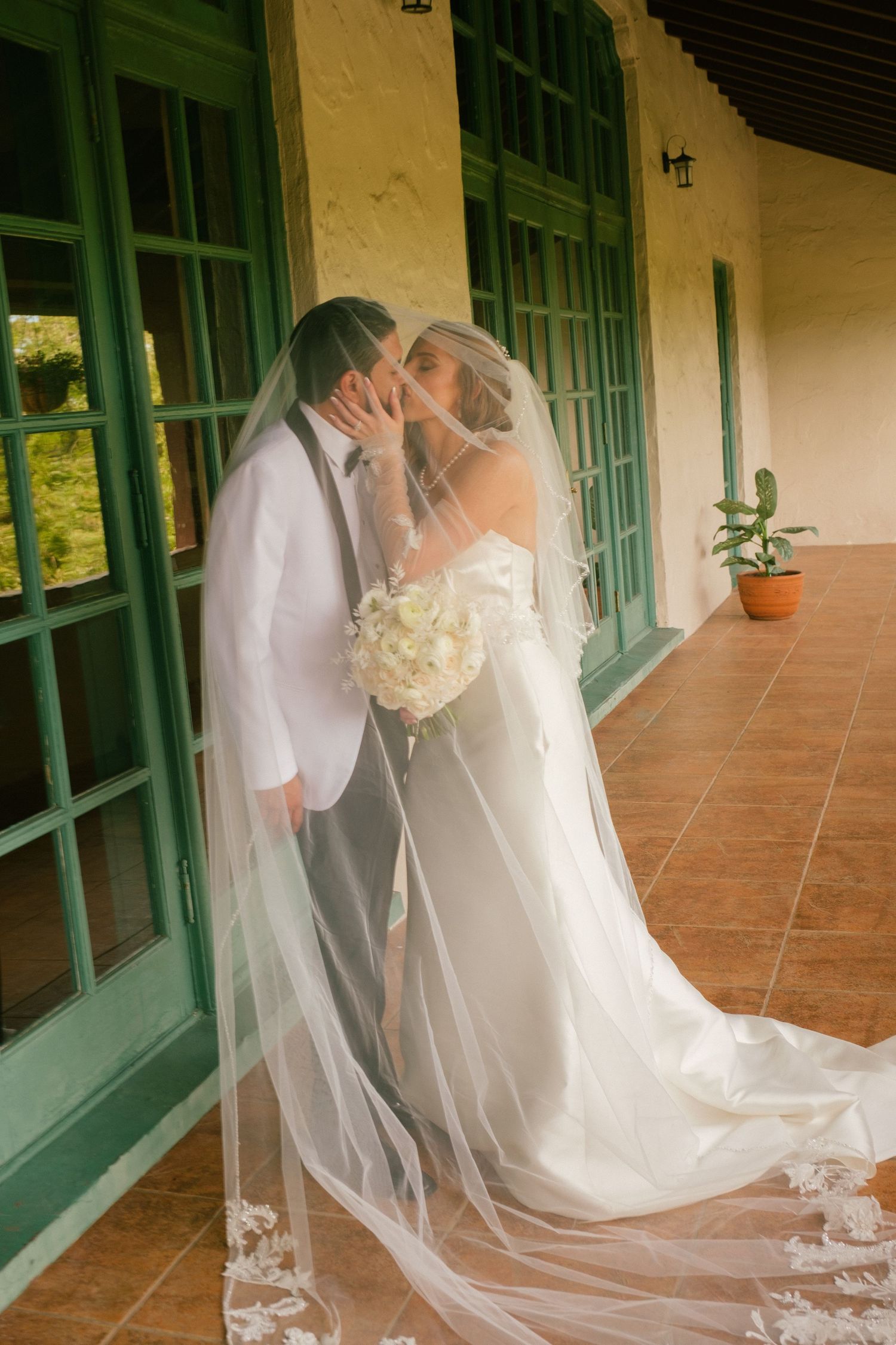 A couple shares a romantic kiss on a porch with green doors and a flowing white veil and wedding dress.