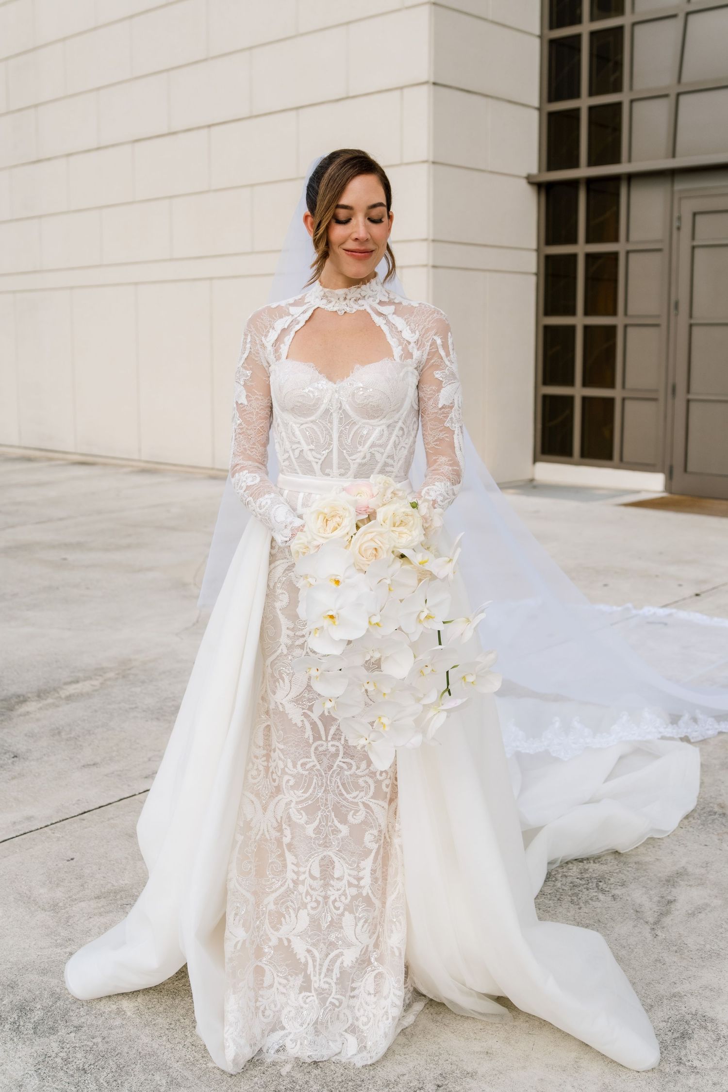 A bride in an elegant lace wedding gown with a flowing veil holds a white orchid bouquet on a light stone terrace.
