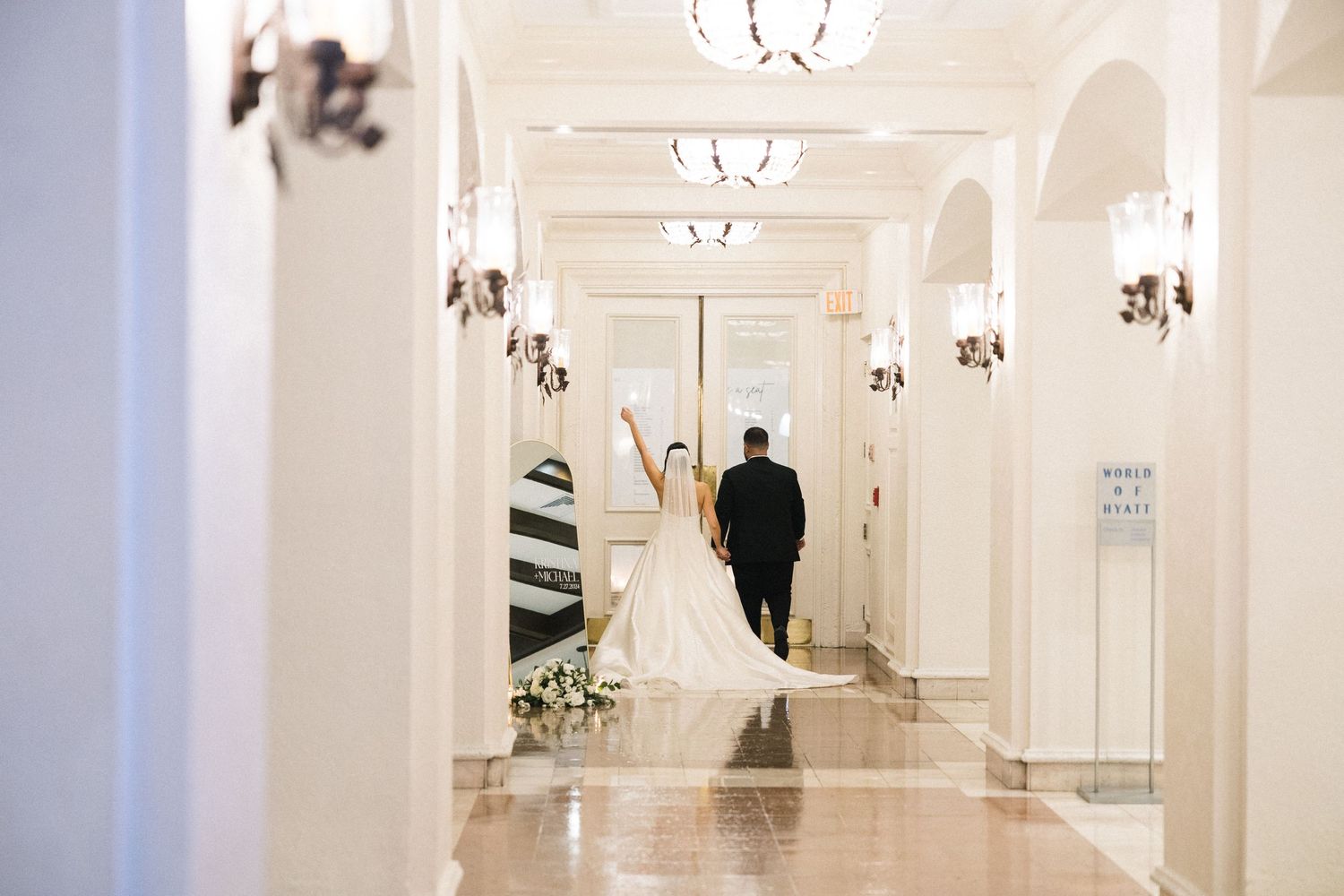 A bride and groom walk down an elegant white hallway with crystal wall sconces in a historic venue.