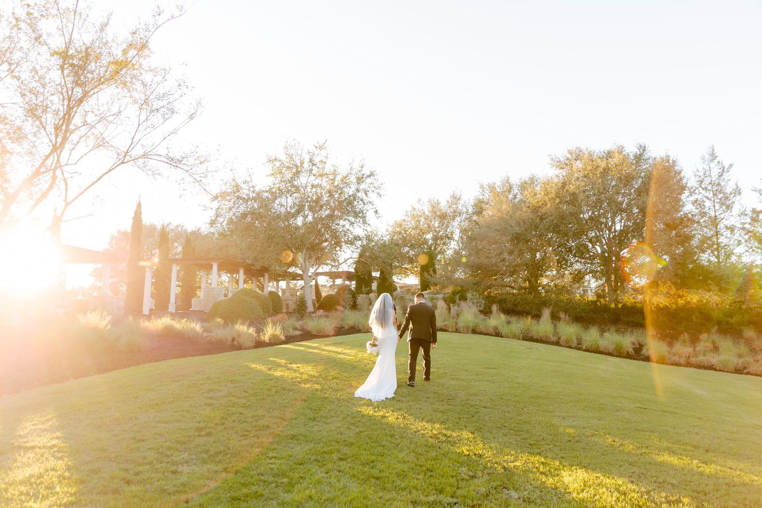 A couple walks on a lush green lawn during golden hour with sun flare and autumn trees in the background.