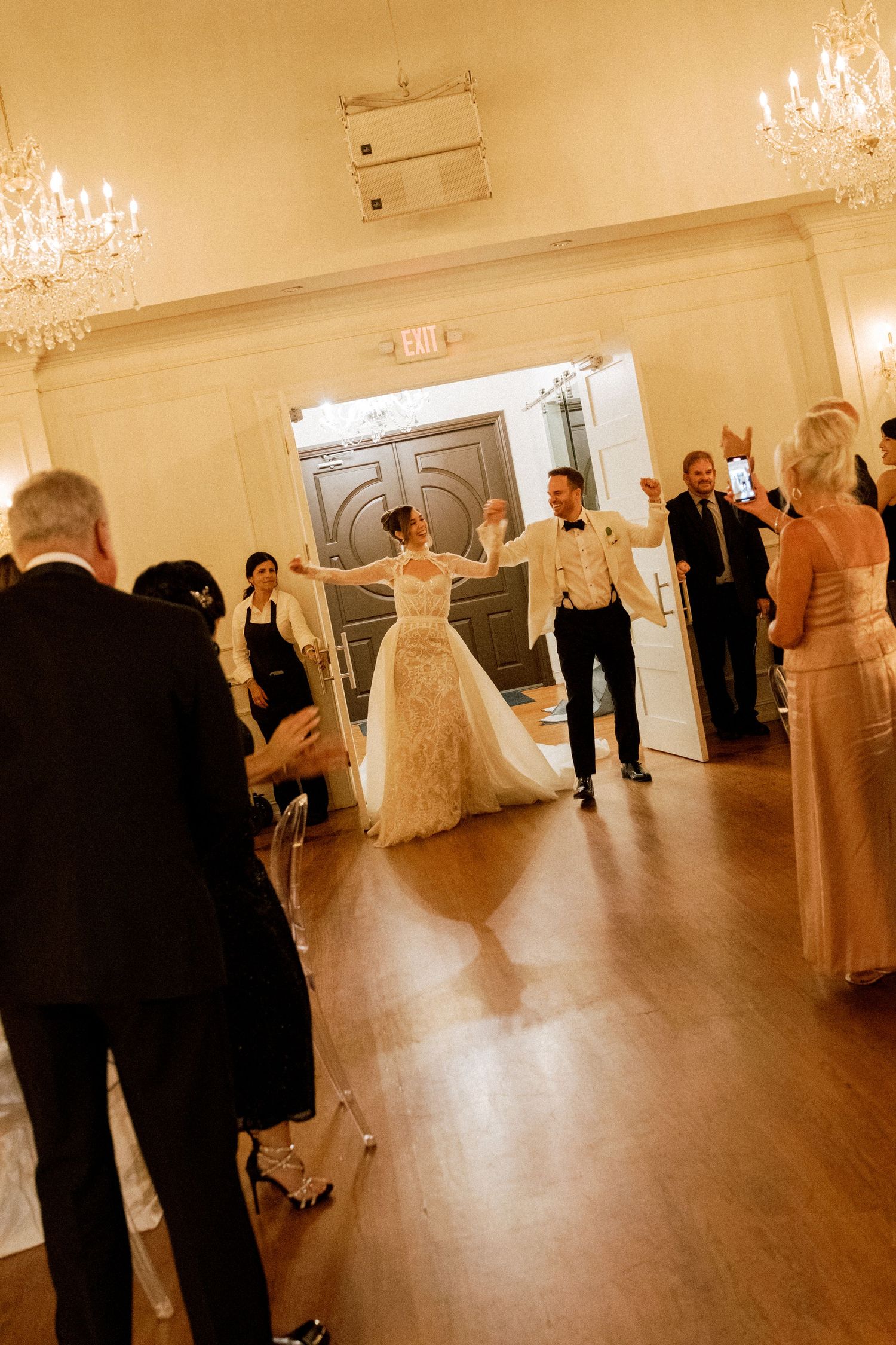 Elegant ballroom wedding reception with couples dancing on wooden floors under crystal chandeliers.