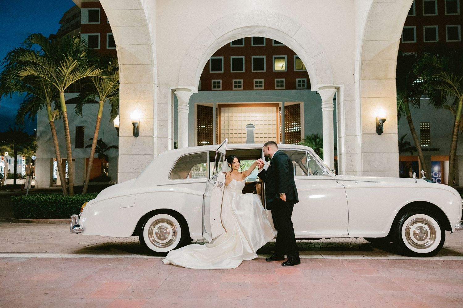 Elegant white vintage Rolls-Royce car parked outside Mediterranean-style building under string lights at evening venue.