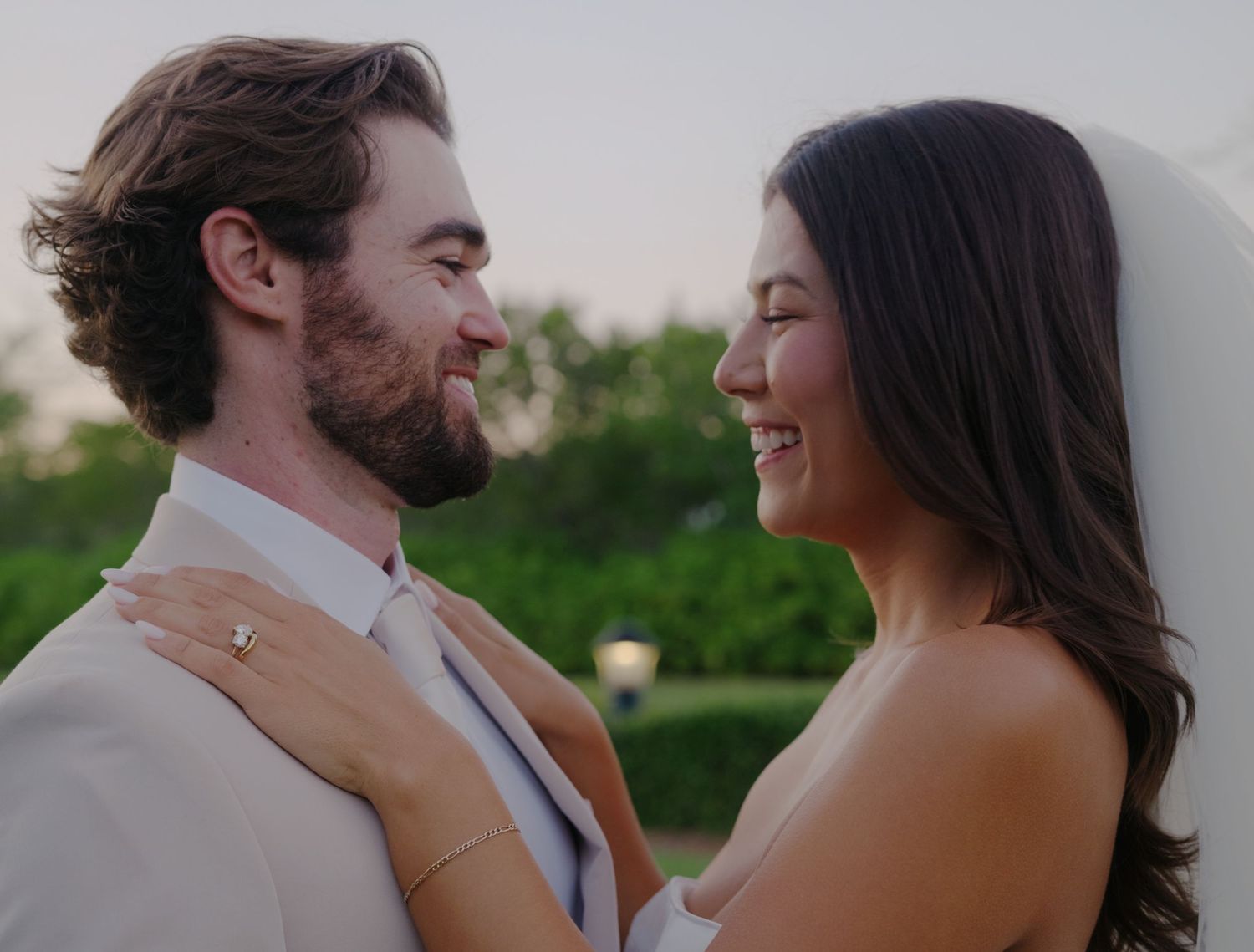 A loving couple sharing a tender moment outdoors, with the bride in white and groom wearing a light beige suit.