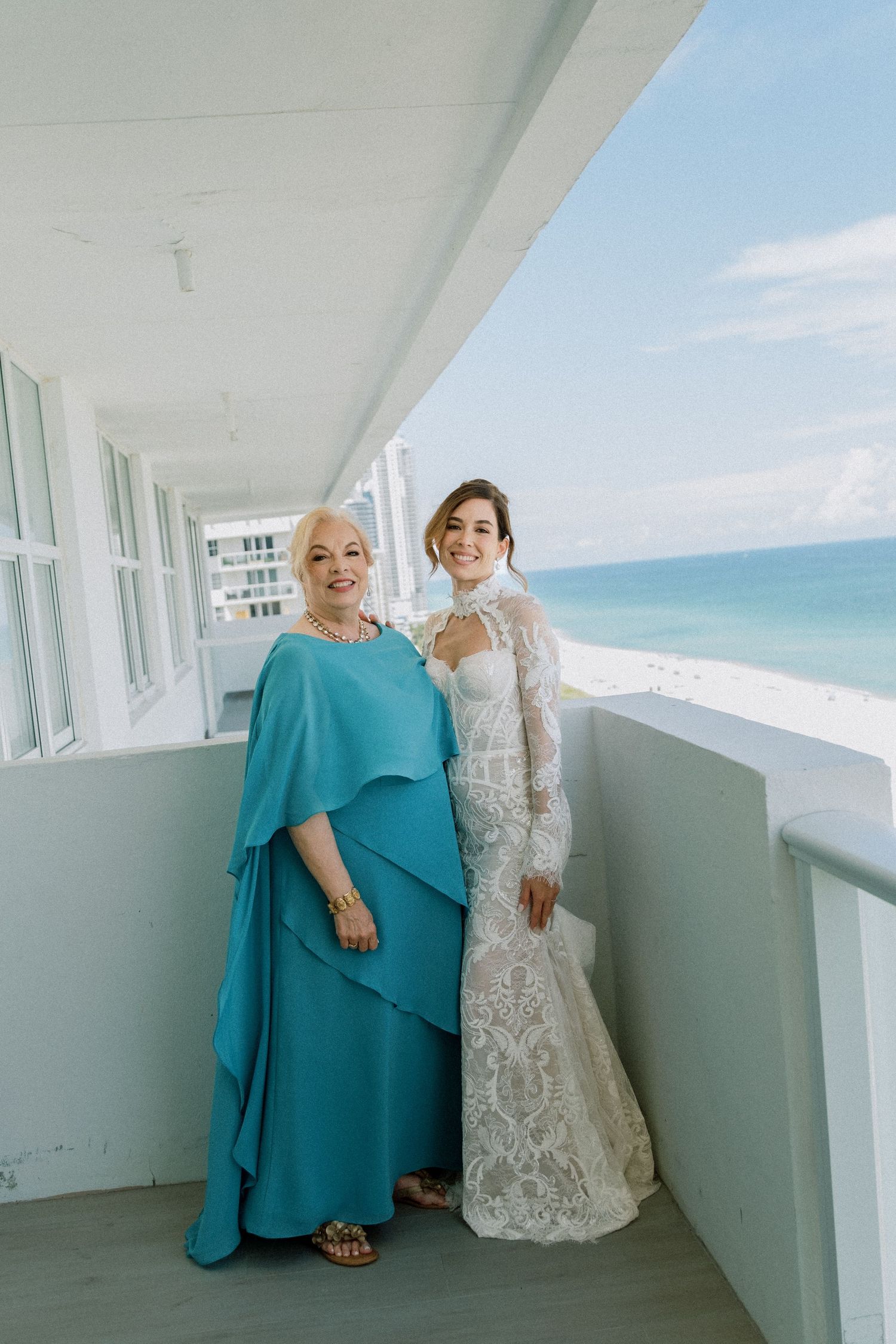 Two people share a tender moment on a white balcony overlooking a bright blue ocean view on a sunny day.