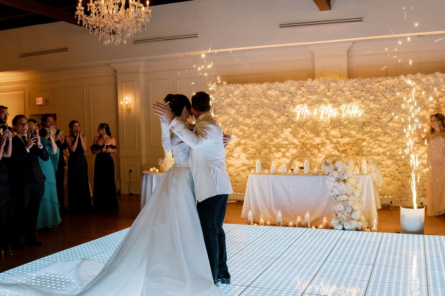 A wedding reception venue features an elegant white dance floor with glowing chandeliers and romantic lighting overhead.