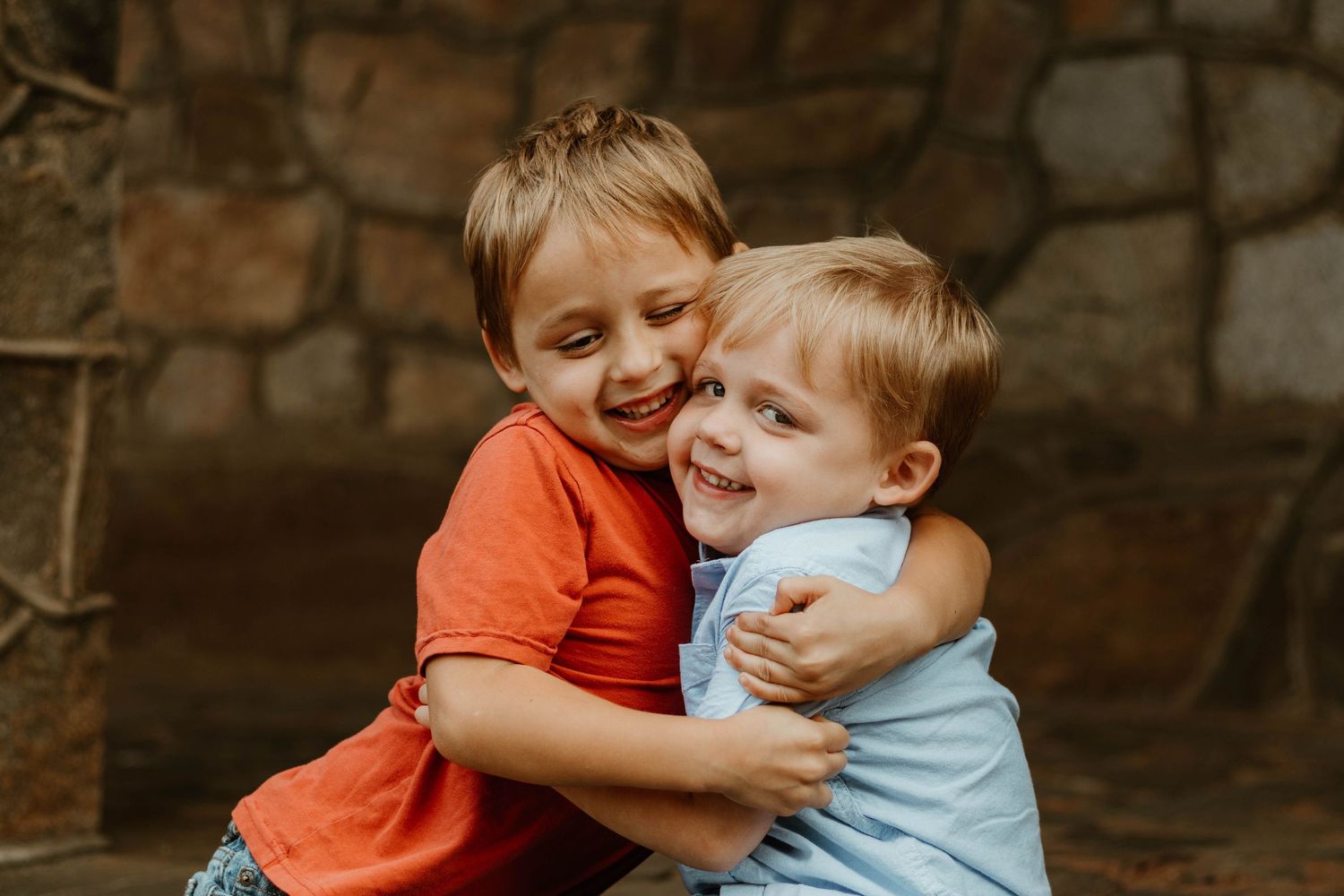 Two children in red and blue shirts share affectionate hugs in front of a rustic stone wall.