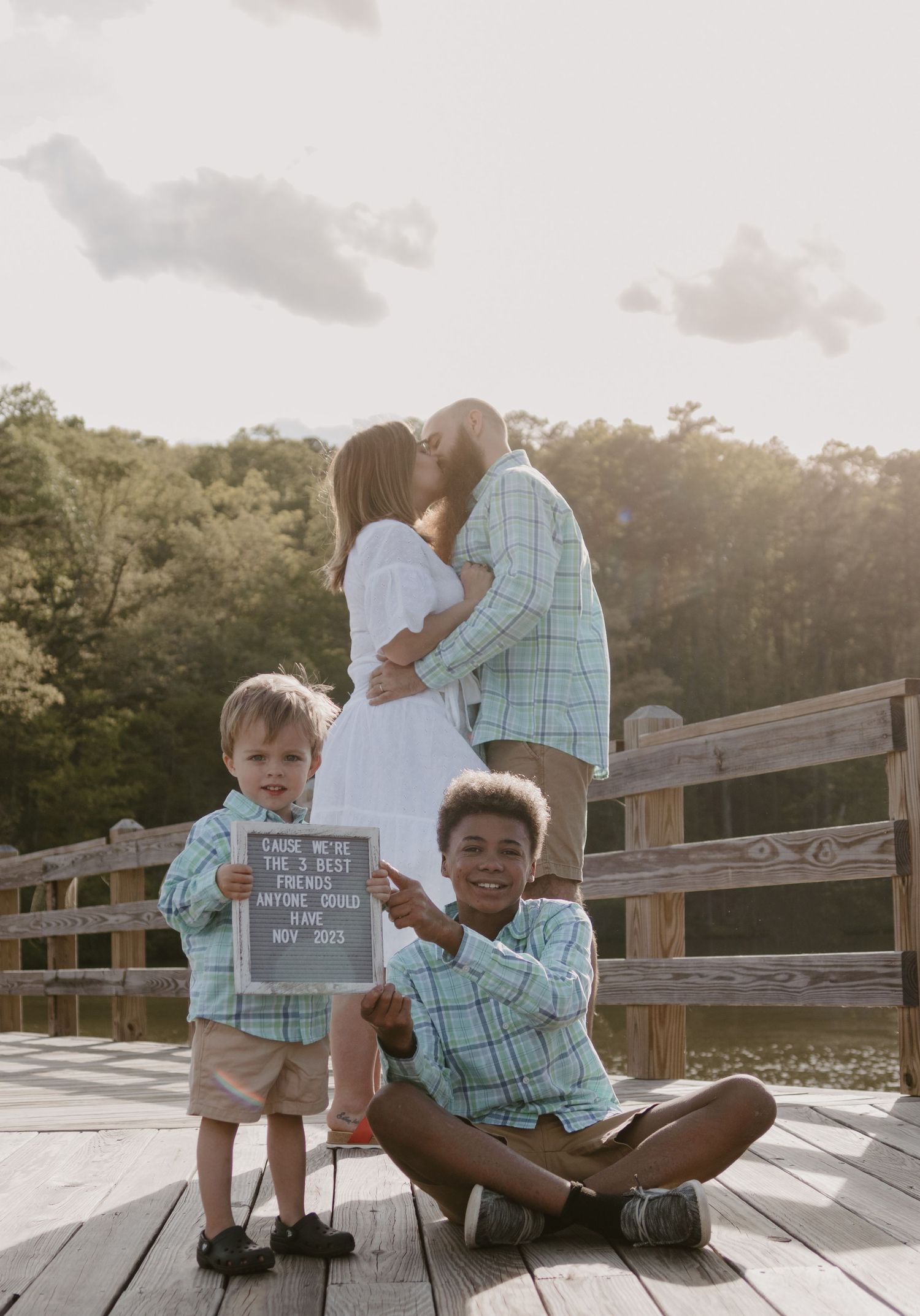 Children holding announcements on a wooden deck while adults embrace in background at Lake Toccoa.