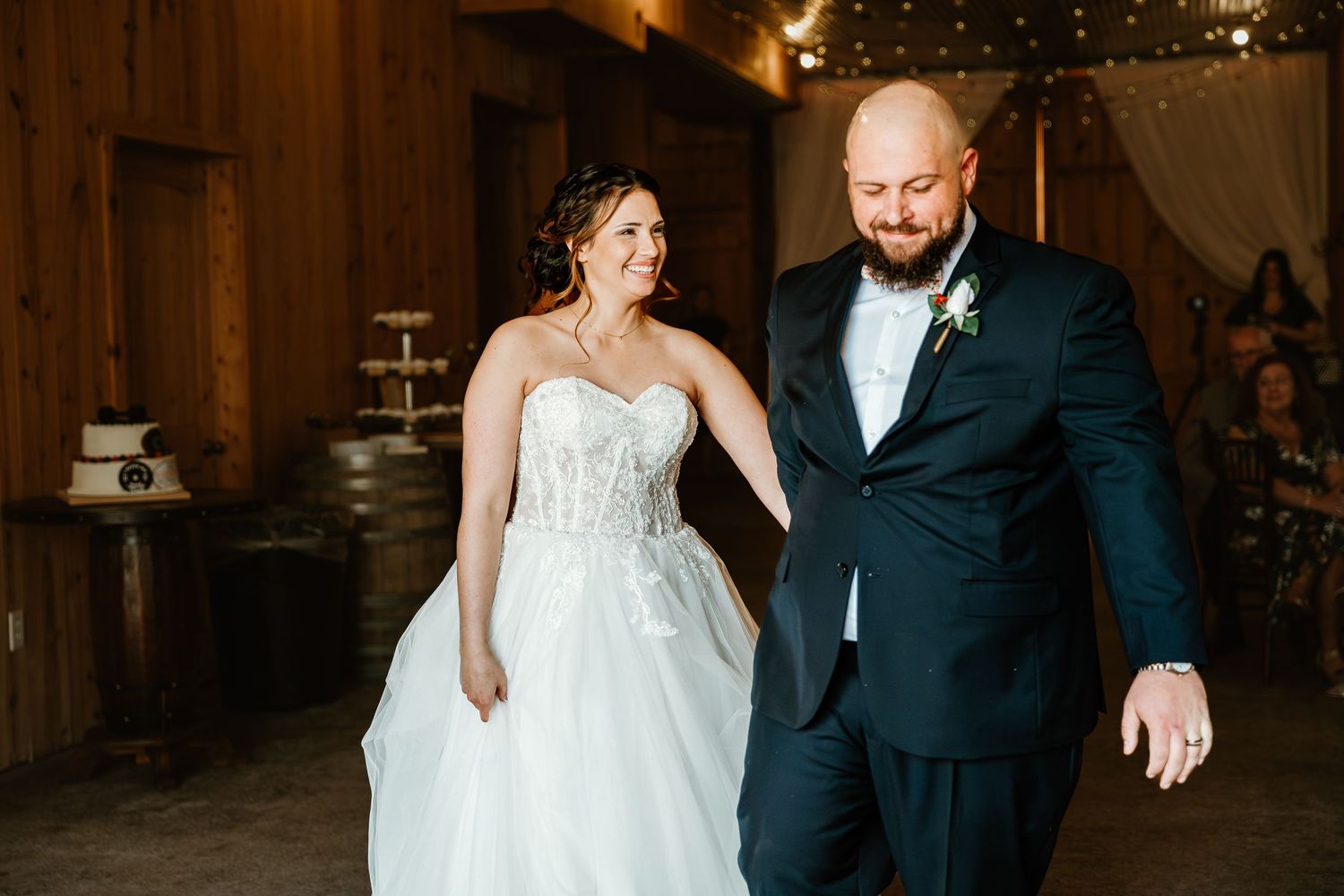 Bride and groom walking hand in hand into rustic barn wedding reception with string lights and cake in background.