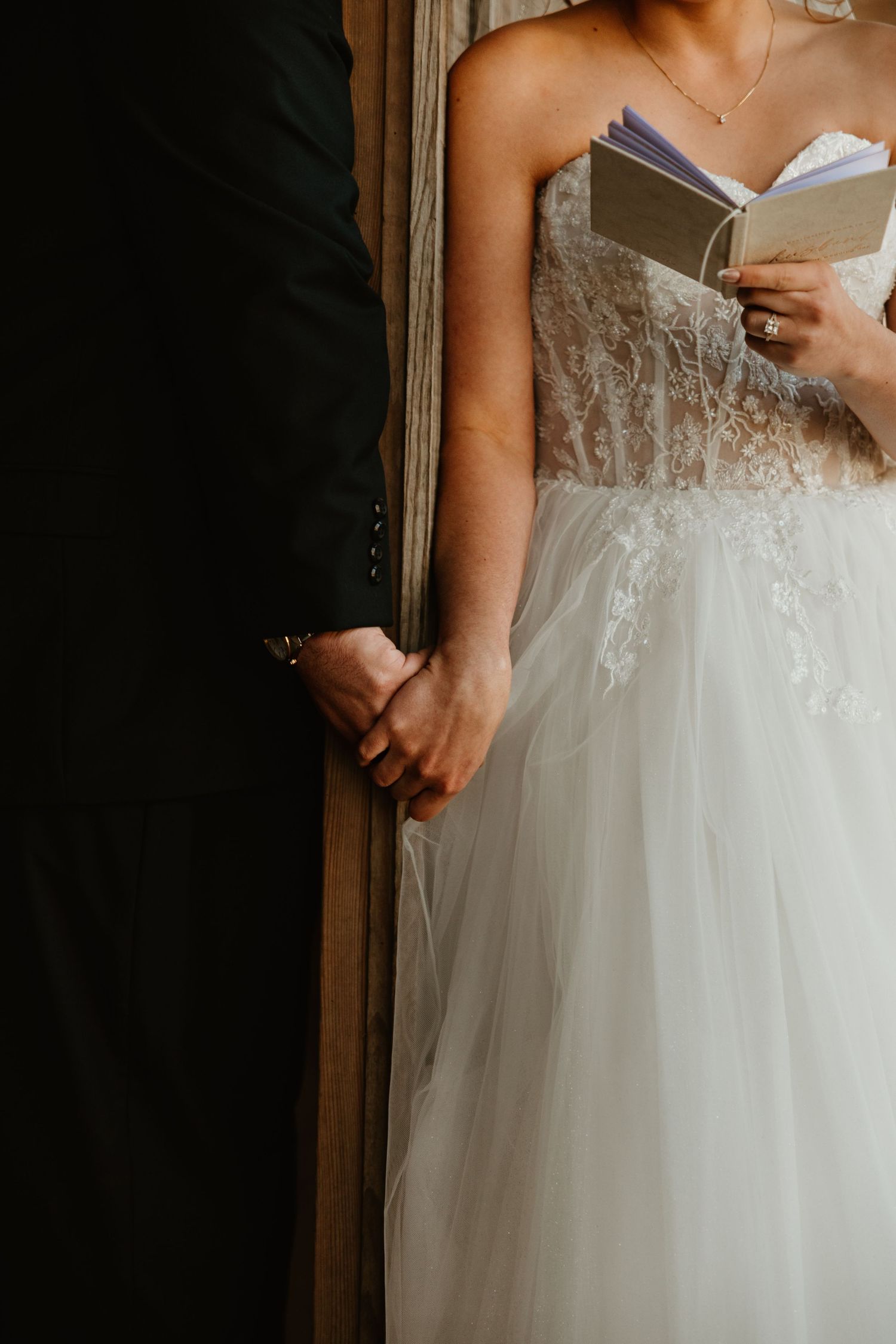 Bride in lace tulle gown holds grooms hand while reading wedding vows from a small book during ceremony.