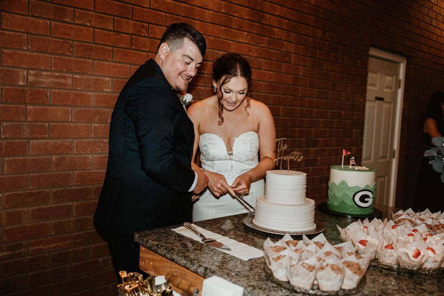 A couple cuts into their elegant white wedding cake together against a rustic brick wall backdrop.