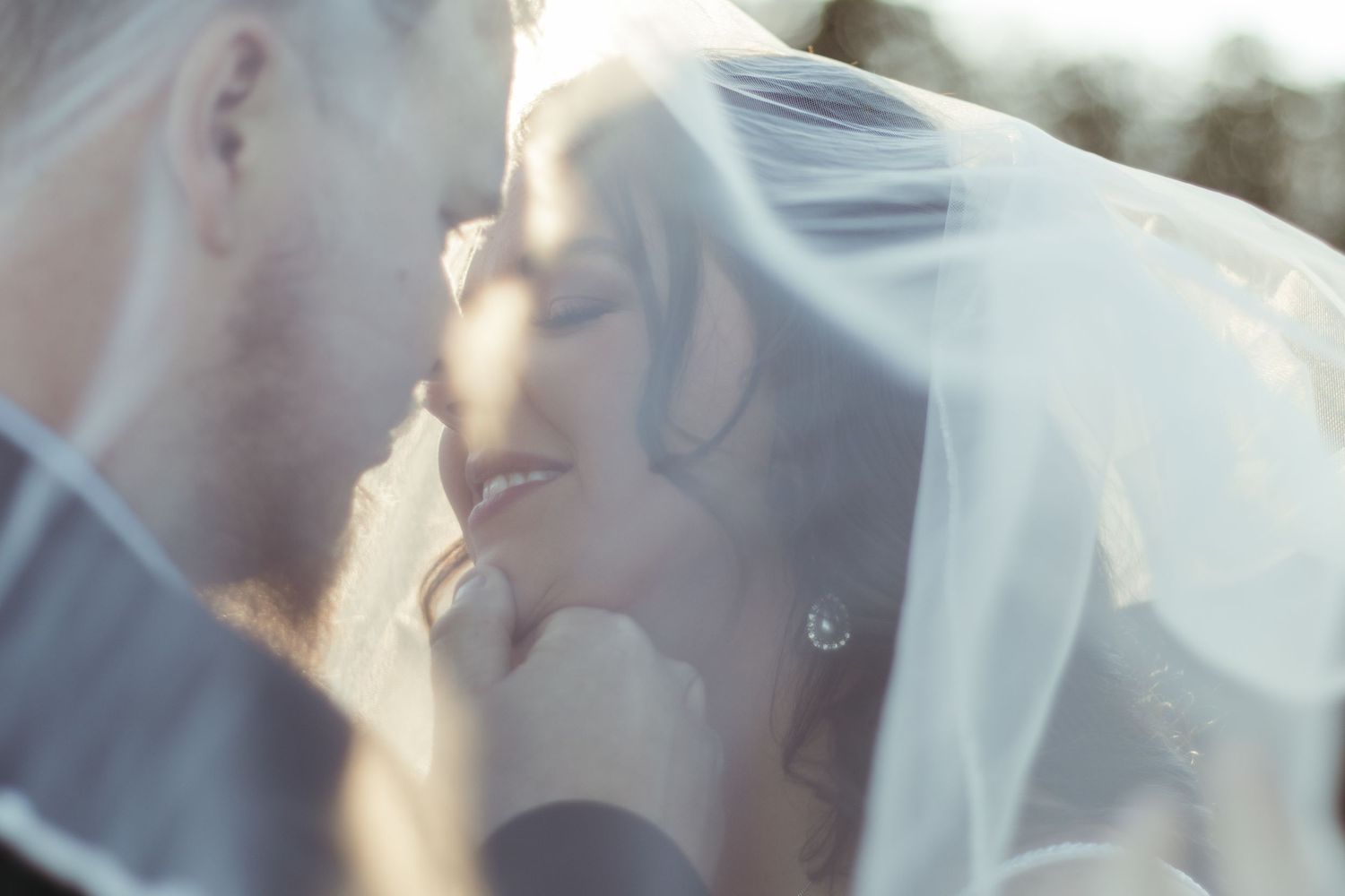 A romantic close-up of a bride and groom sharing an intimate moment under a flowing veil in soft lighting.