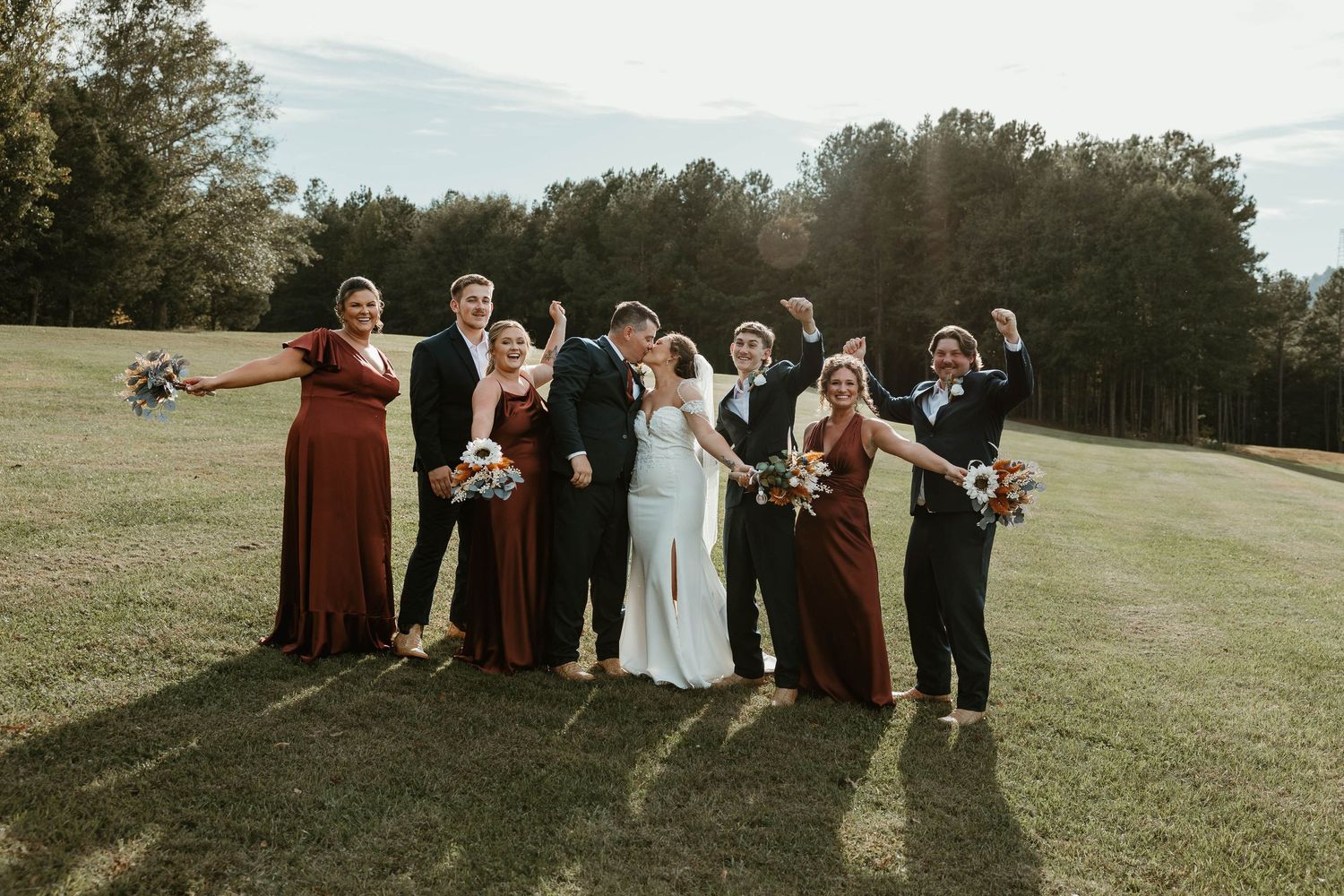 A wedding party stands together on a grassy field with bridesmaids in burgundy dresses on a sunny autumn day.