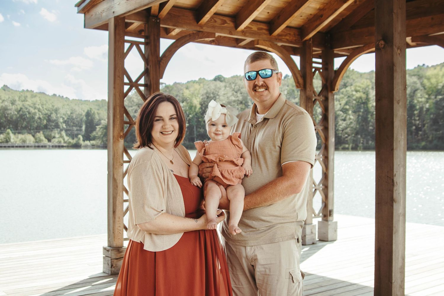 Family wearing coordinated beige and white outfits poses together under a wooden gazebo near a lake.