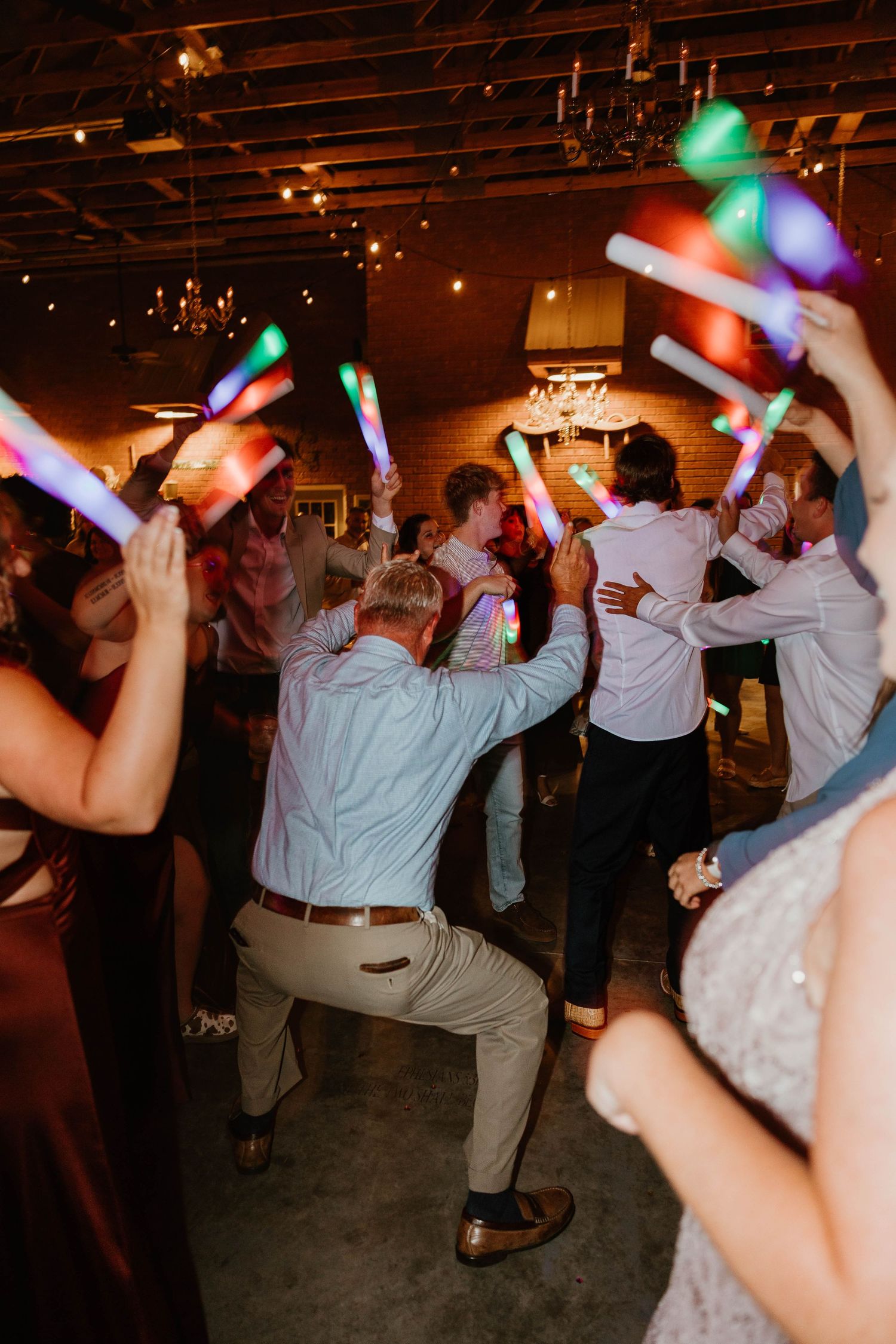 Wedding reception guests dance with colorful light-up foam sticks on a dimly lit dance floor.