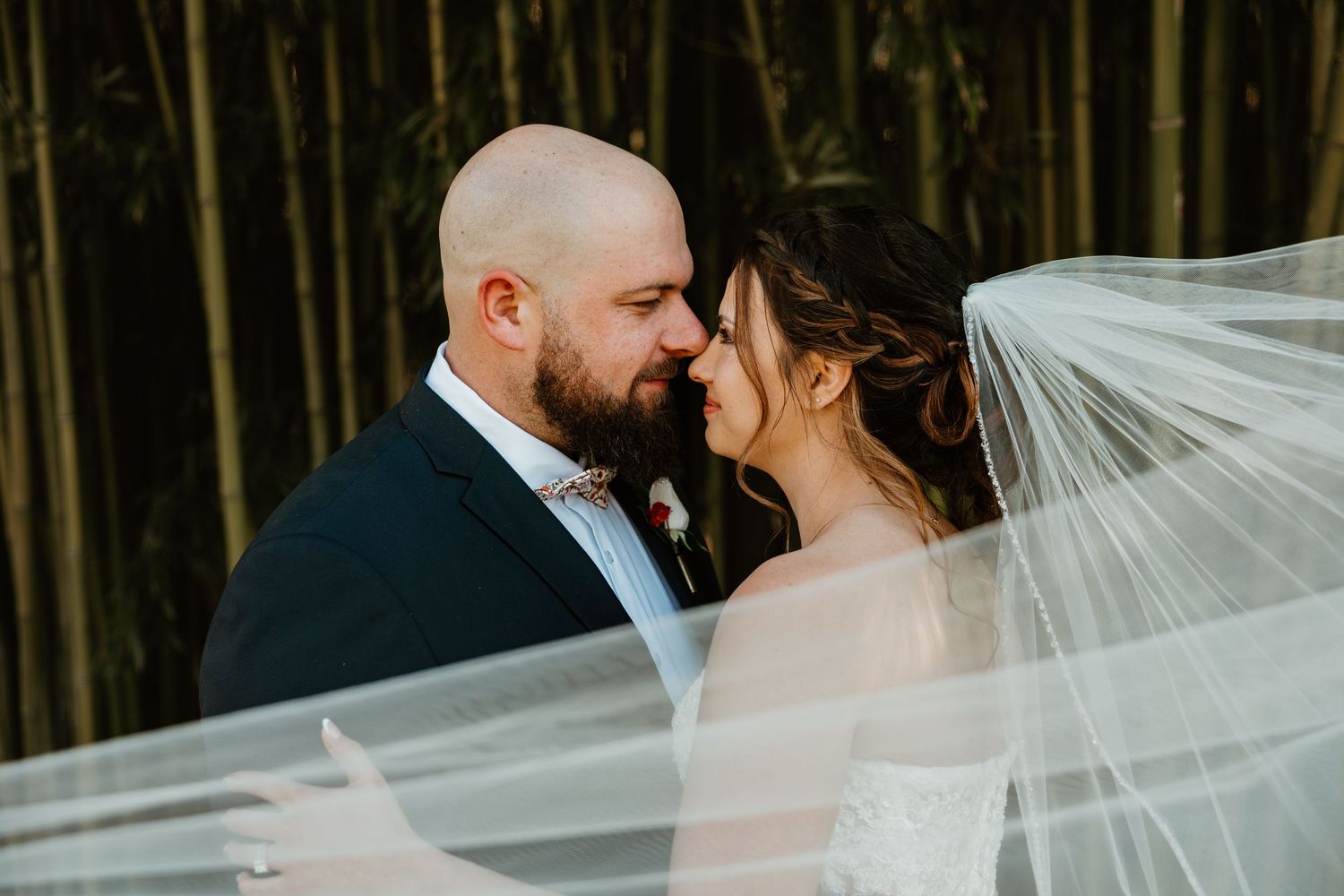 Bride and groom share a romantic moment, veil flowing, with bamboo forest backdrop.