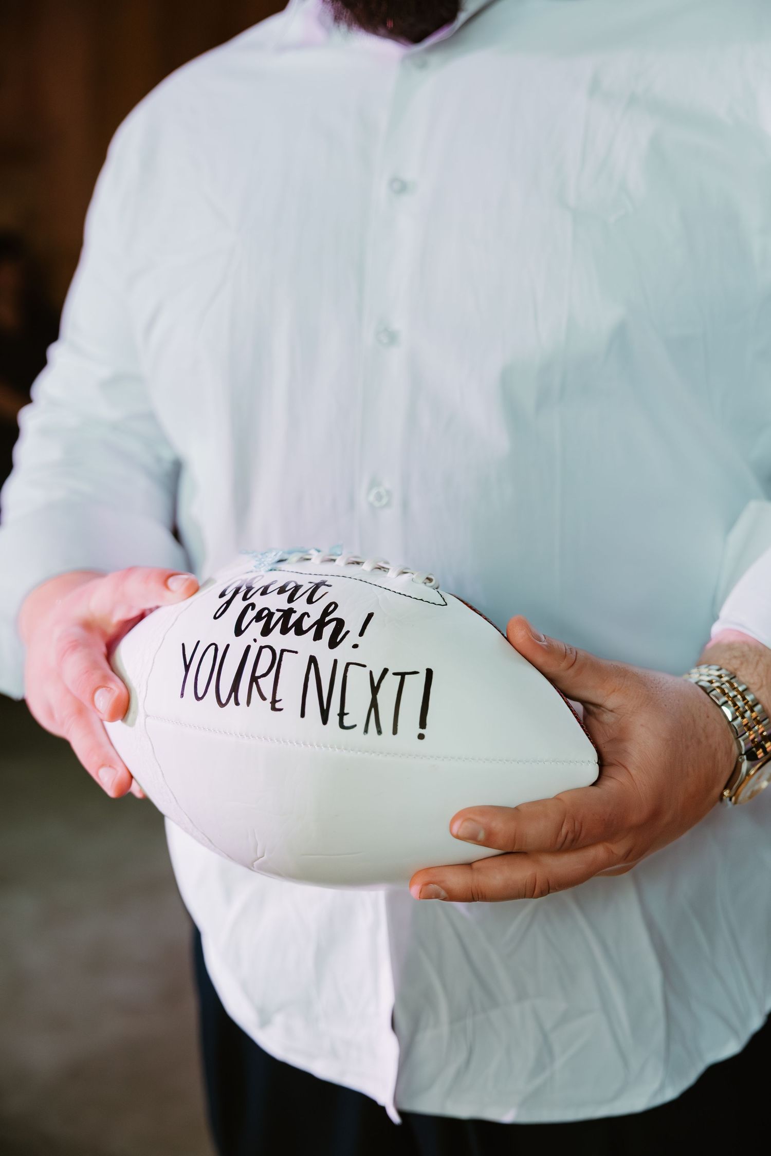 Man in light blue shirt holds white football inscribed with you're next at a wedding reception.