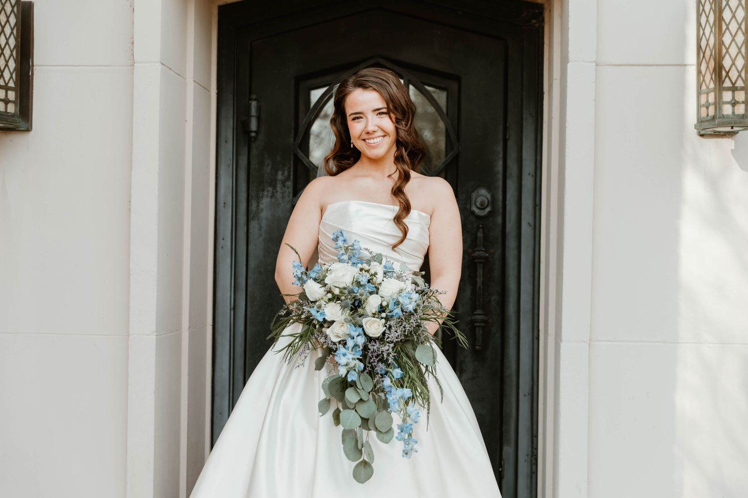 Bride in strapless white ball gown holding cascading blue and white floral bouquet with greenery, standing before dark door.