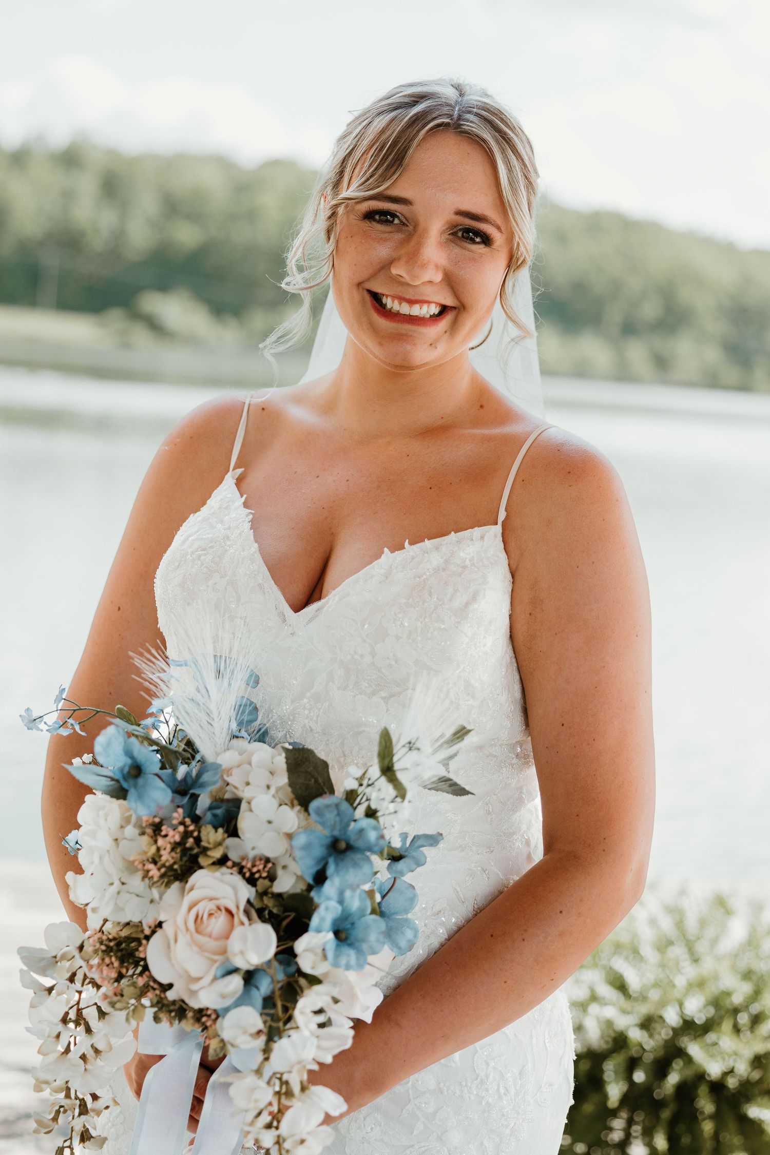 Smiling bride in white lace gown holds blue and white floral bouquet outdoors near a lake.