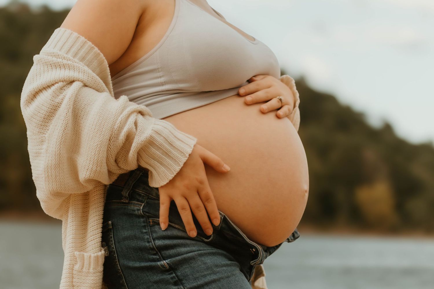 Side view of pregnant belly being gently held while wearing cream cardigan and jeans by Lake Hartwell.