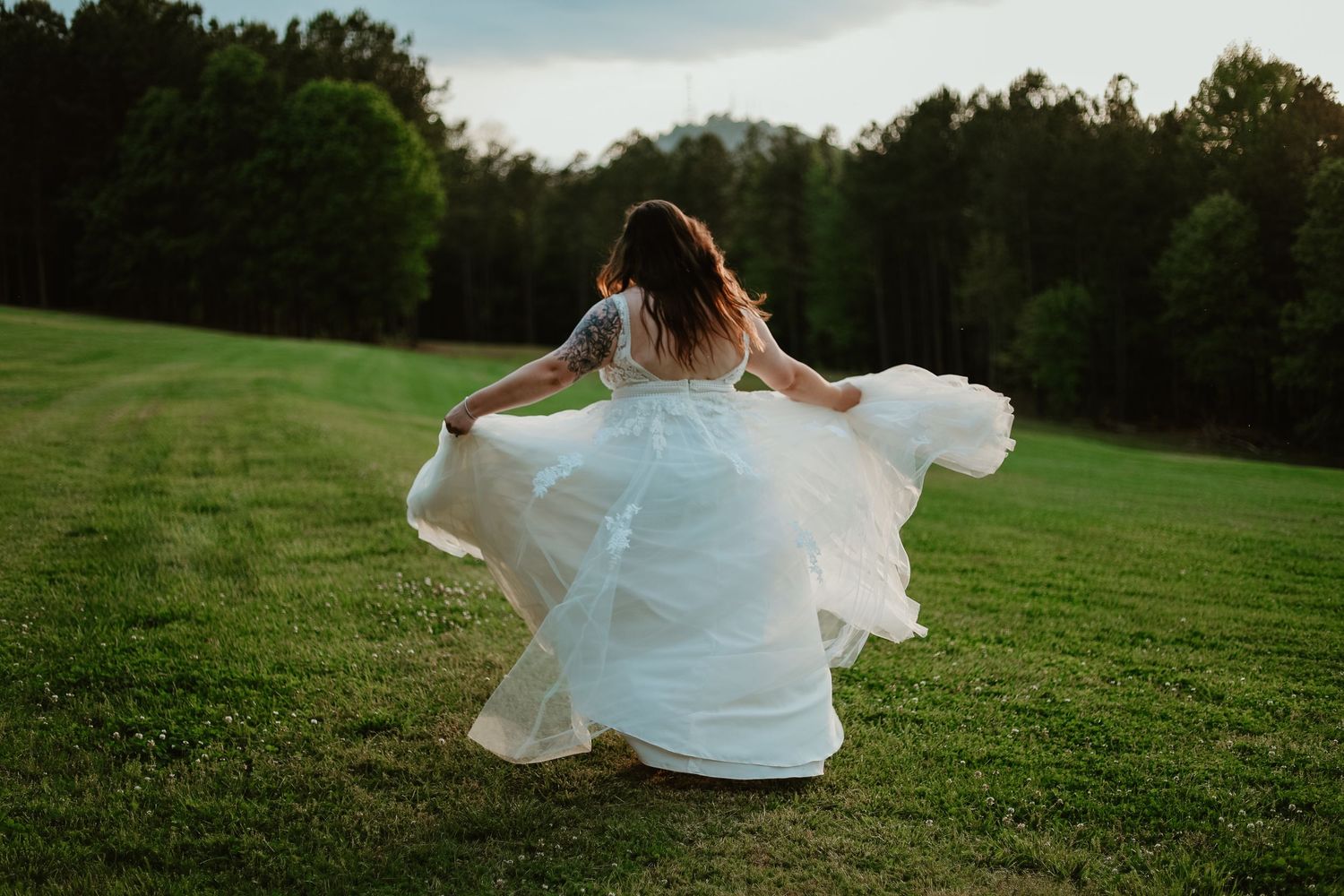 A person in a flowing white wedding dress twirls gracefully across a lush green hilltop meadow at sunset.