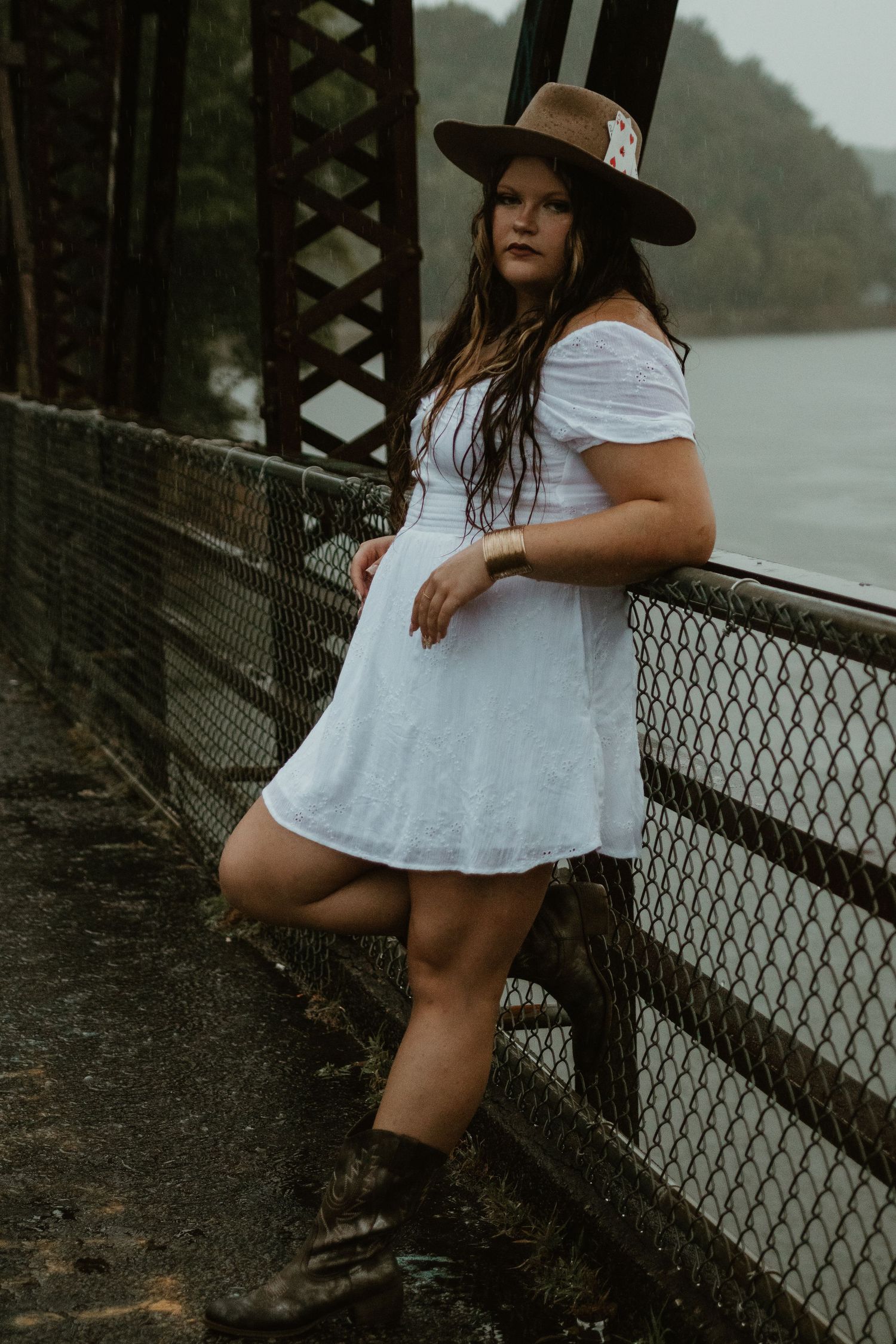 Person in white dress poses against chain link fence on Broken Bridges on overcast day.