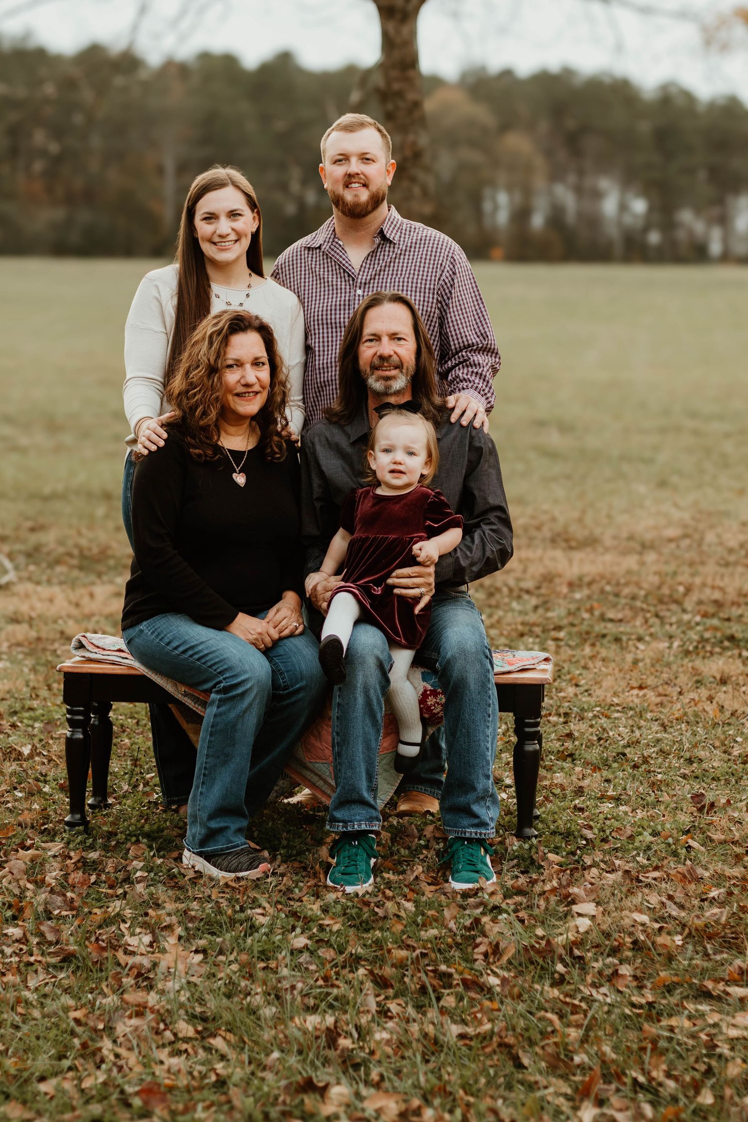 Family poses together on wooden bench surrounded by fallen autumn leaves in outdoor setting in Commerce, GA.