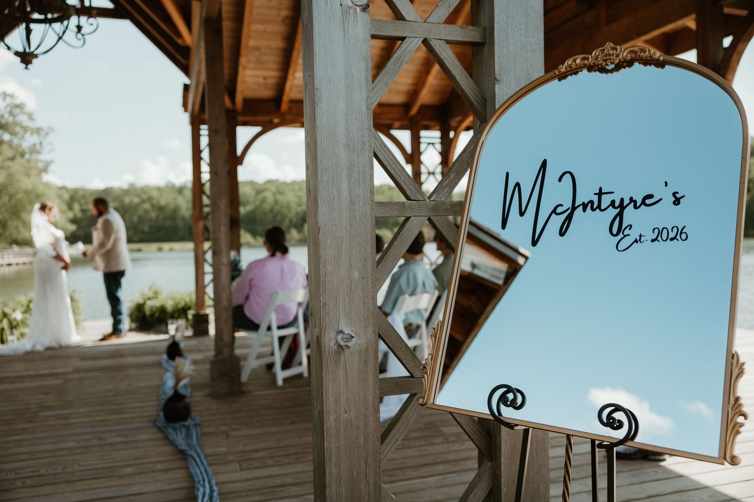 Outdoor wedding ceremony on wooden pavilion with McIntyre's Est. 2026 mirror sign in foreground near lake.