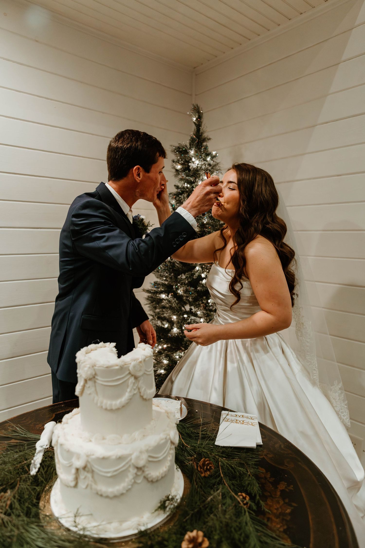 Newlyweds share intimate moments while cutting their white wedding cake decorated with evergreen branches.