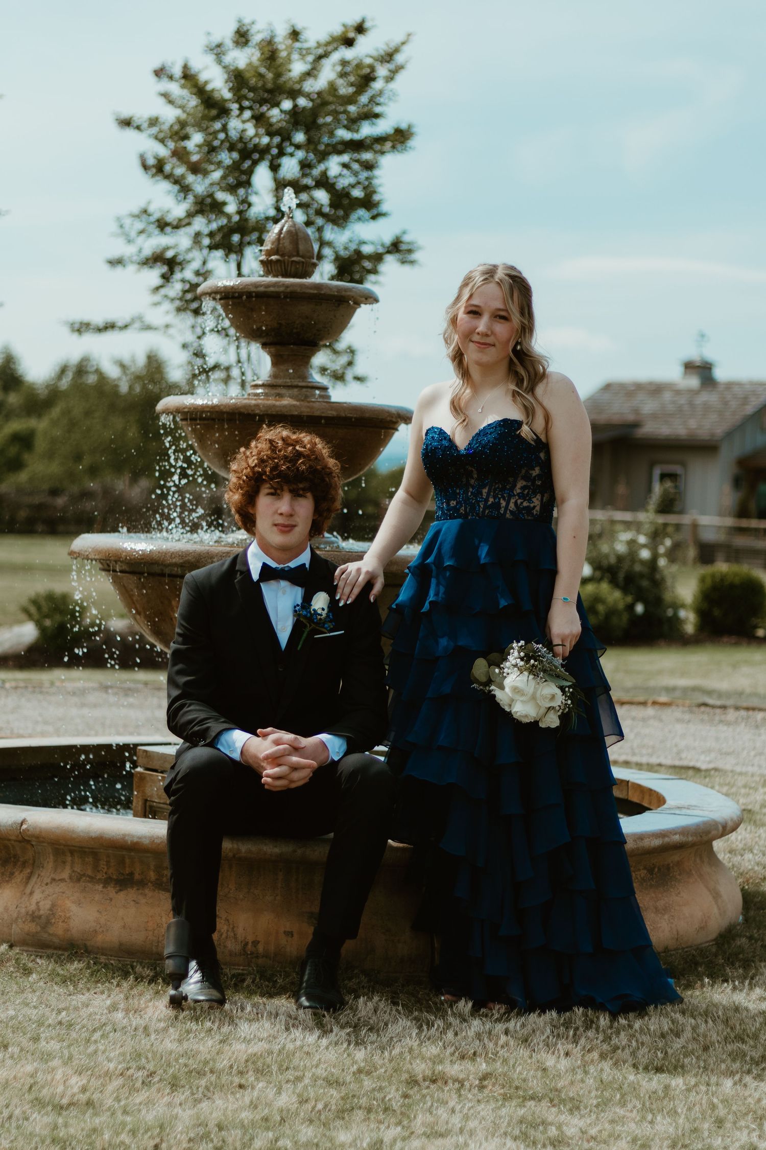 A formal couple poses by an elegant fountain in blue gown and black tuxedo at The Venue at Broken Bridges in Toccoa, GA.
