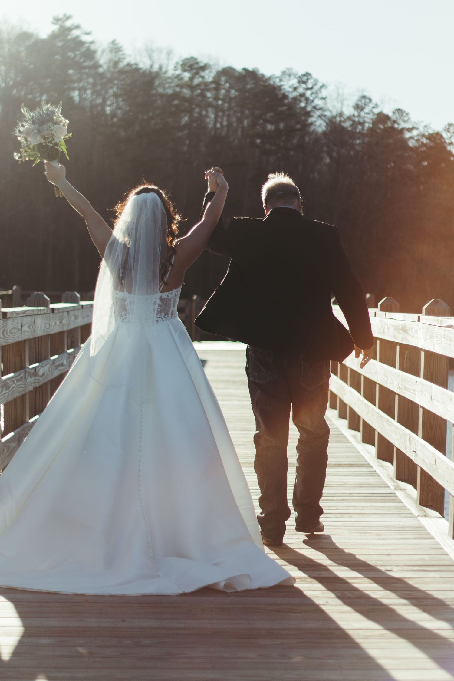 A bride in a flowing white dress and groom stand together on a wooden boardwalk at sunset creating silhouettes.