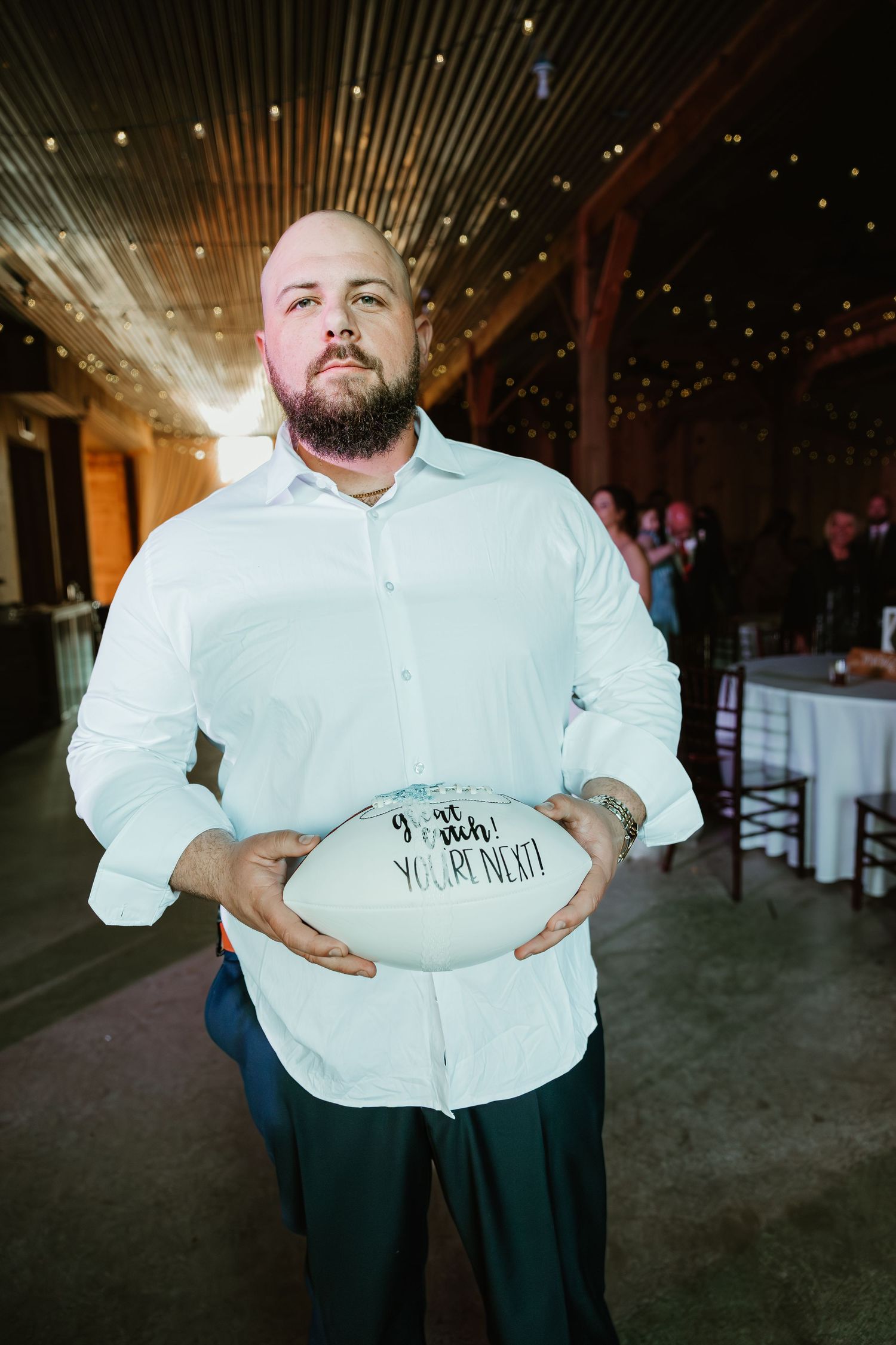 Bald bearded man in white shirt holds football signed I got picked youre next at rustic barn wedding reception.