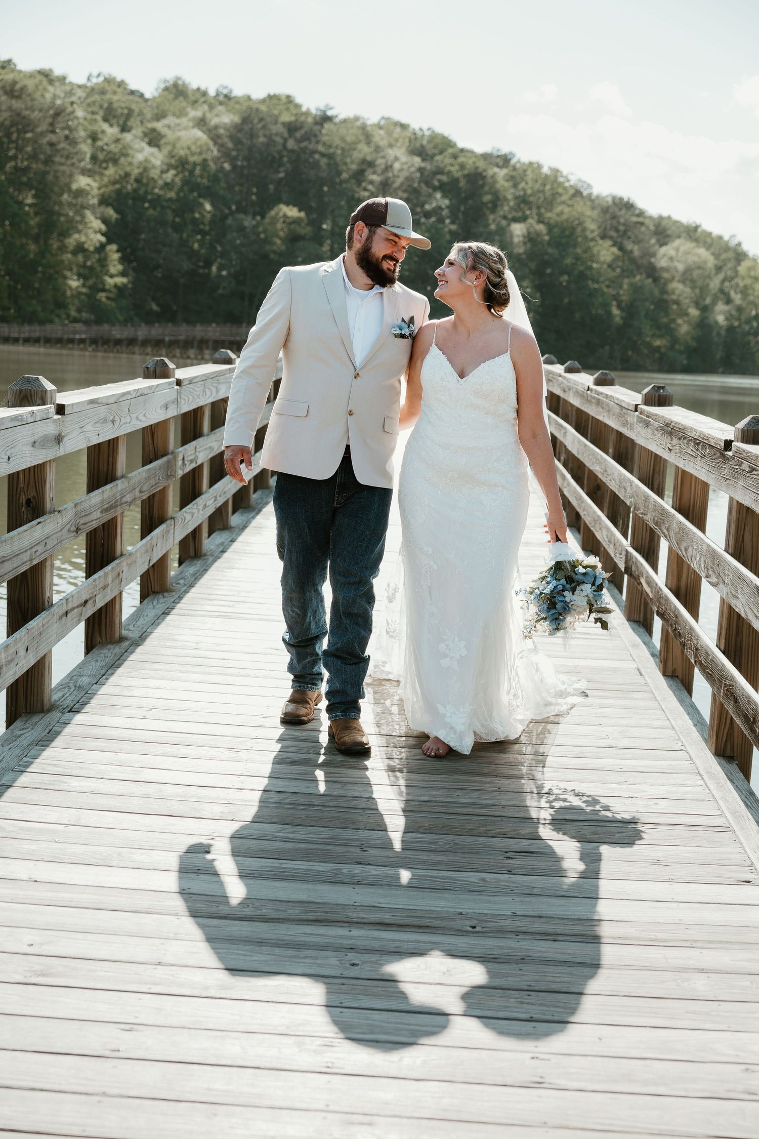Bride and groom walking hand-in-hand on a wooden dock, smiling at each other, with lush trees and water in the background.